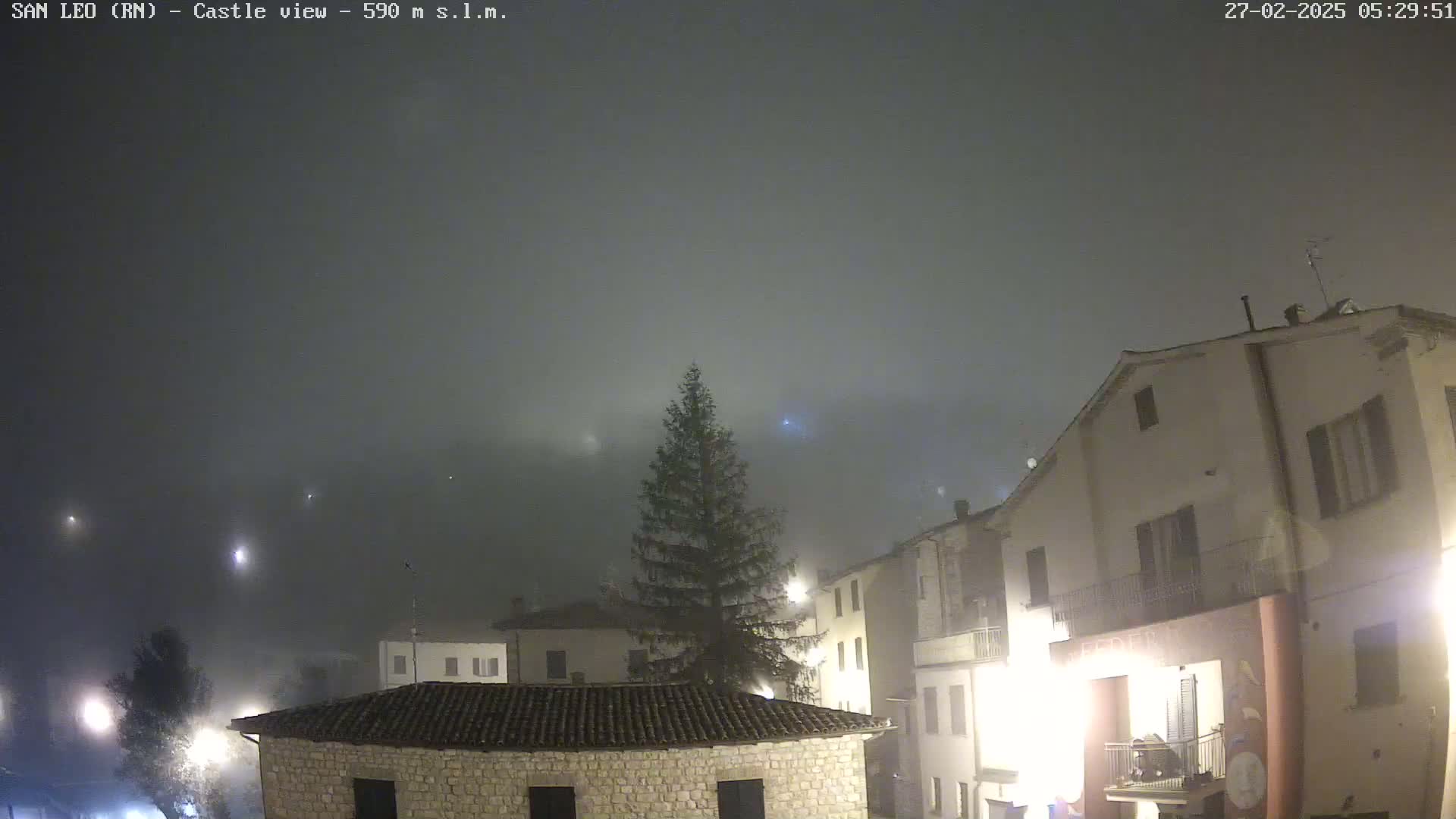 A foggy night scene shows a village of stone buildings illuminated by streetlights, with a tall evergreen tree in the center.