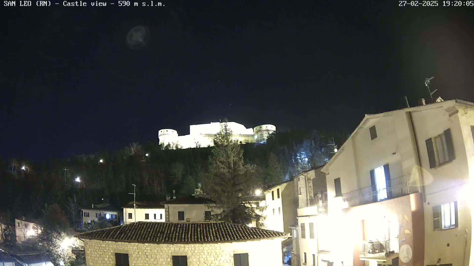 A brightly lit castle sits atop a hill overlooking a small town at night under a clear, dark sky.