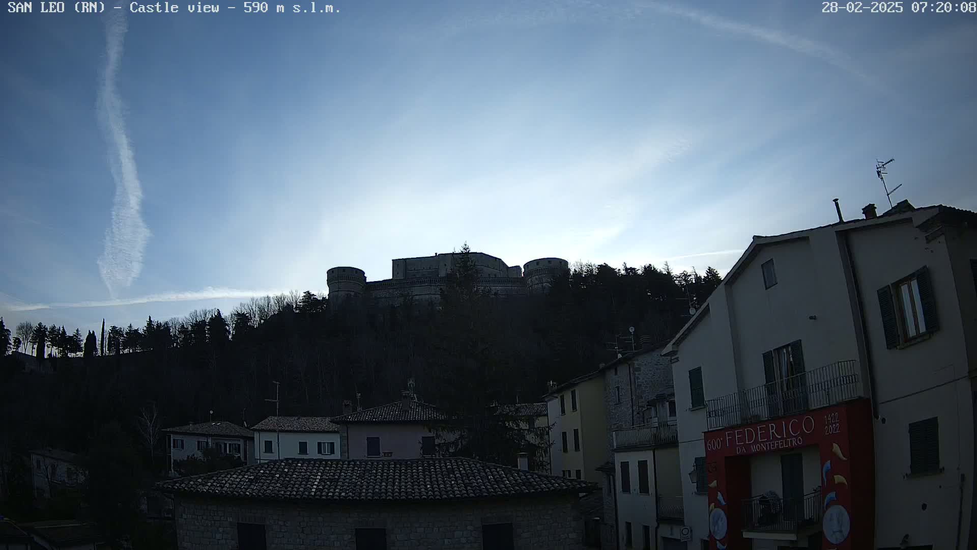 A large stone castle sits atop a hill overlooking a town of buildings under a clear, twilight sky with a contrail.