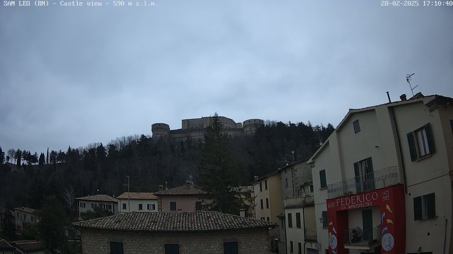 A stone castle sits atop a hill covered in leafless trees under a cloudy sky, overlooking a town of stone buildings with terracotta roofs.