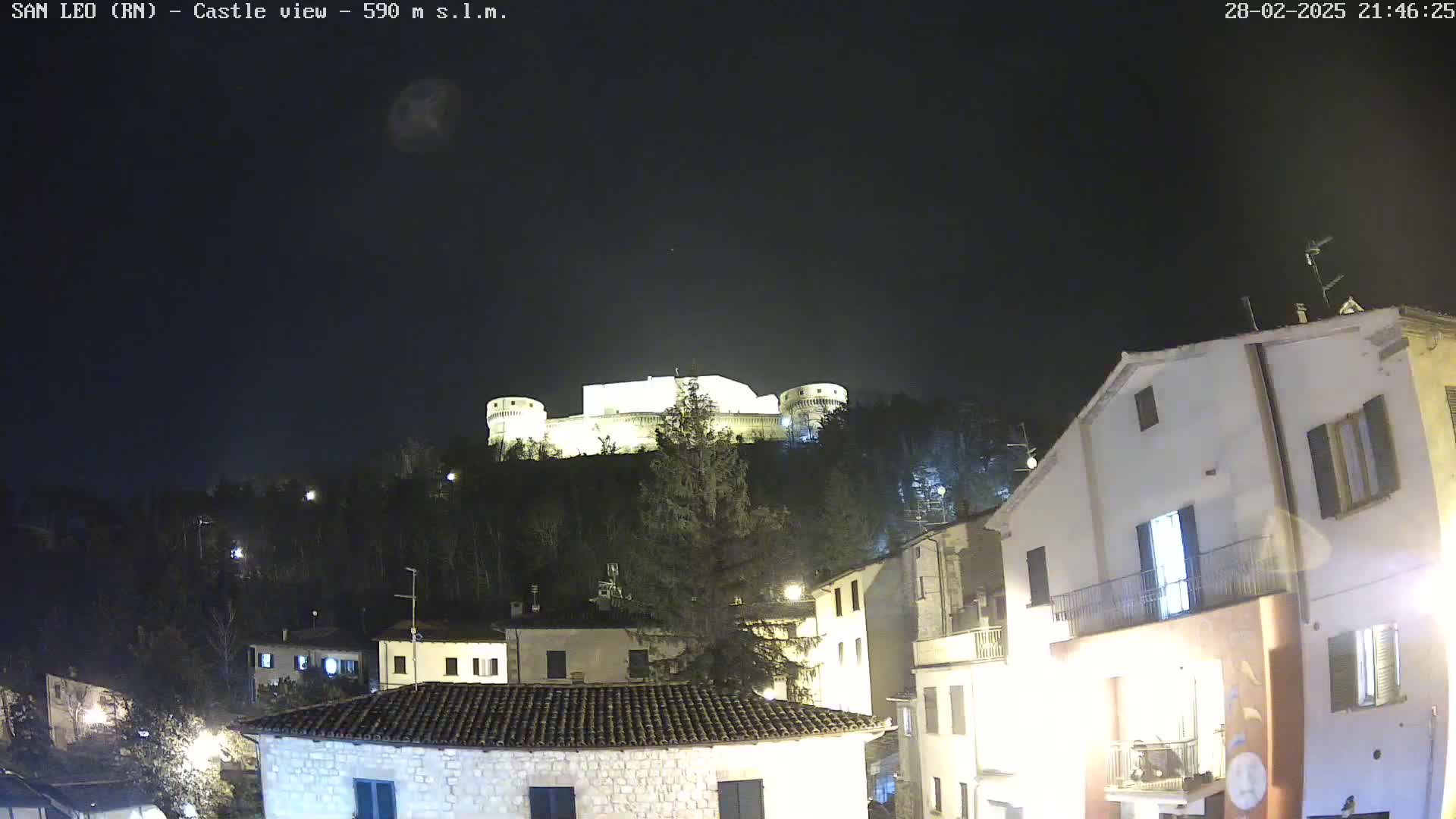A nighttime view of a hilltop castle illuminated against a dark, clear sky, overlooking a village with buildings lit up.