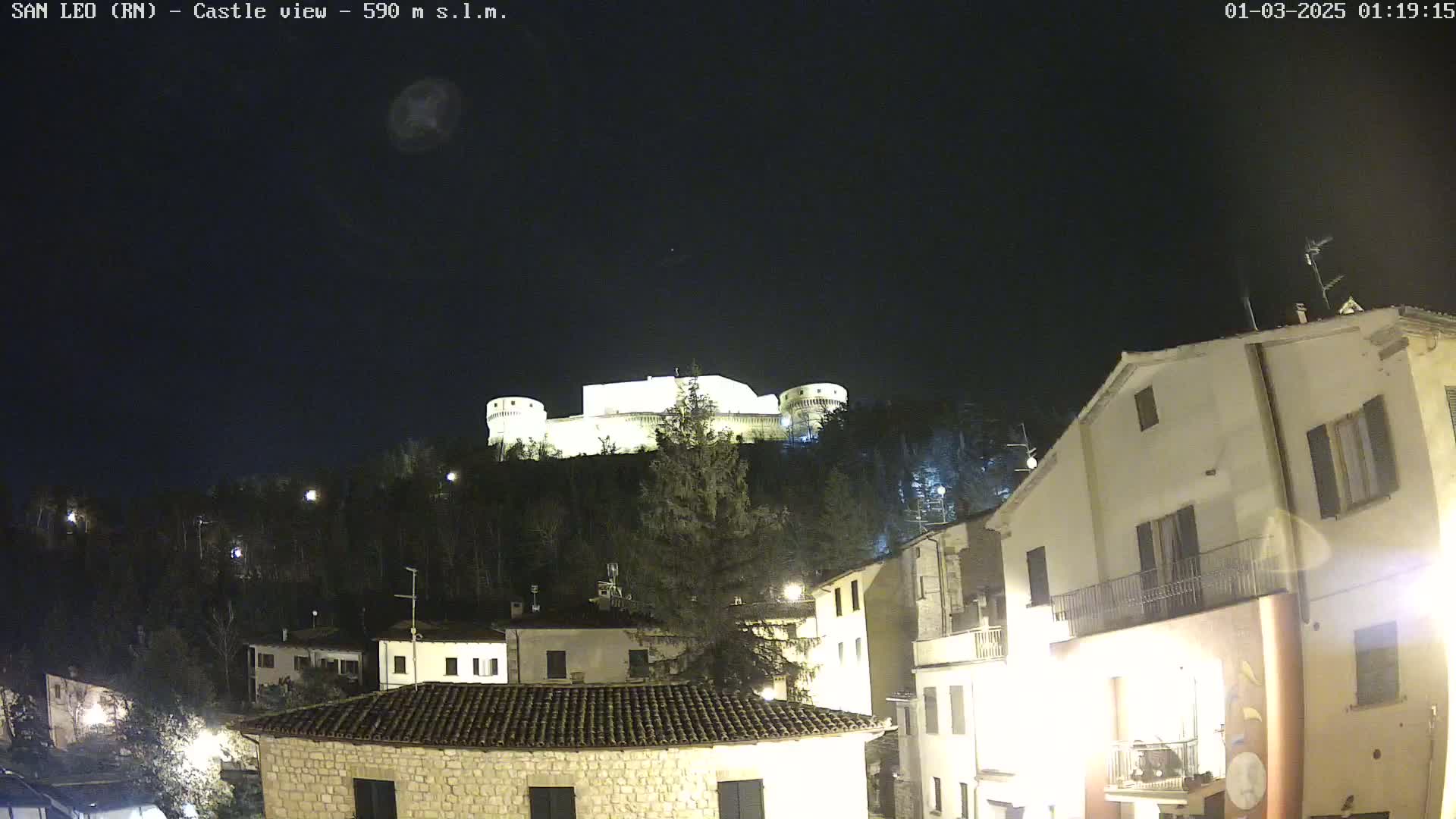 A nighttime view of a hilltop castle illuminated against a dark, clear sky, overlooking a village with buildings lit up below.