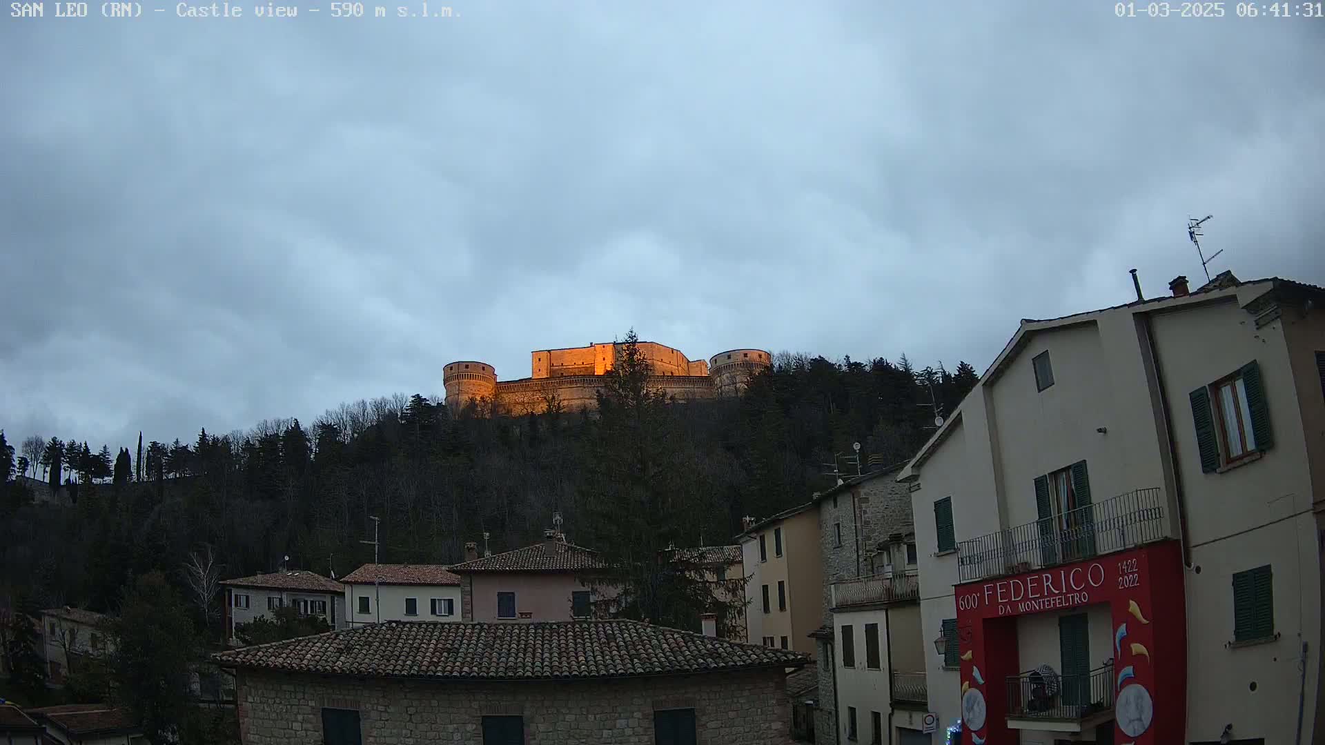 A stone castle sits atop a hill, illuminated by the low morning sun, overlooking a town of closely clustered buildings under an overcast sky.