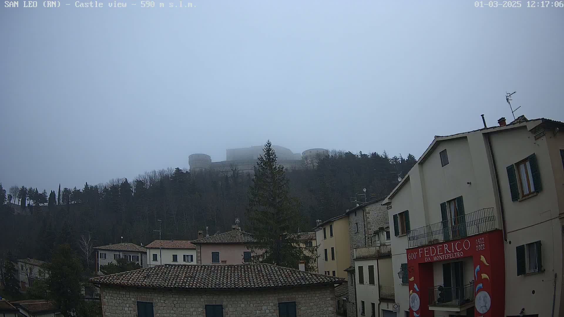 A foggy, overcast day reveals a hilltop castle overlooking a town of closely-spaced buildings with terracotta roofs.