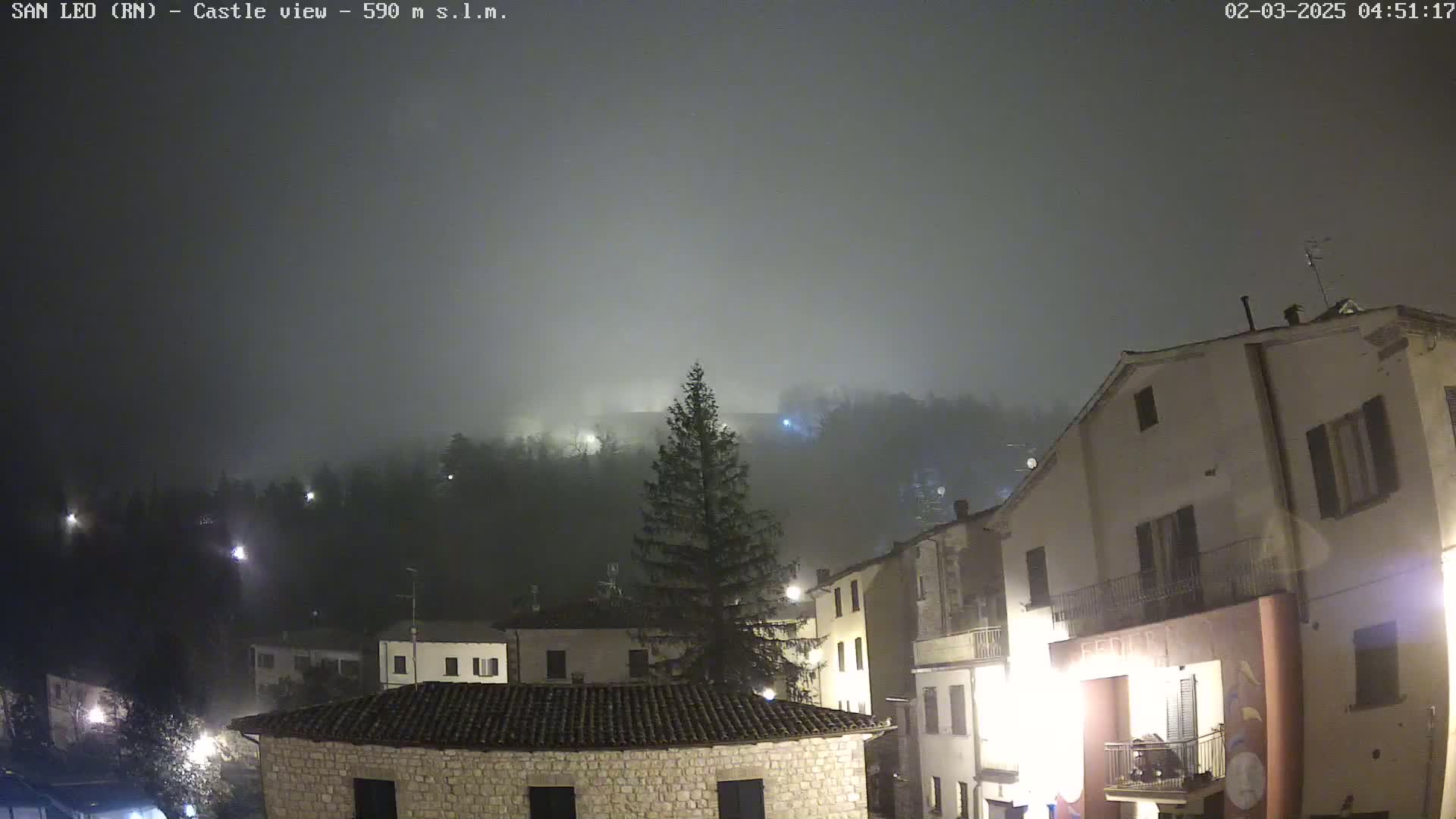 A foggy night scene shows a village of stone buildings illuminated by streetlights, with a hill covered in fog in the background.