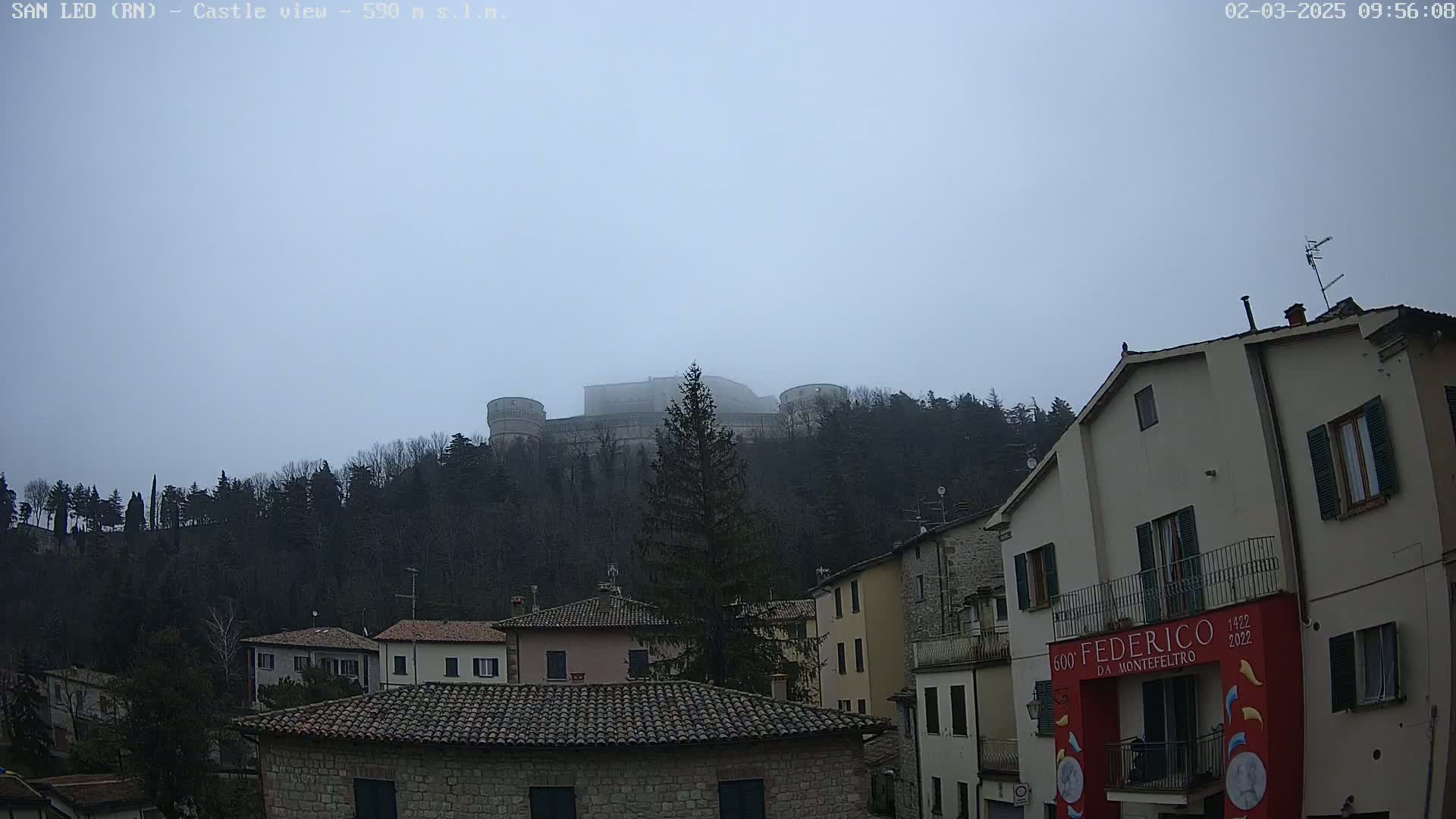 A stone castle sits atop a fog-covered hill behind a village of low stone buildings on an overcast day.