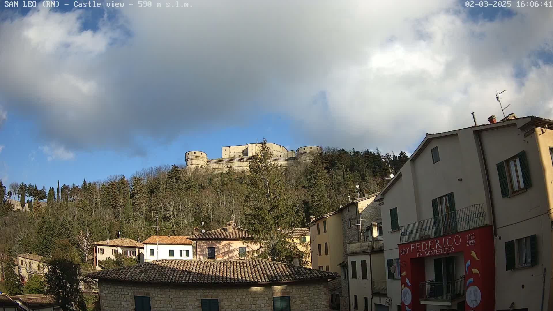 A large stone castle sits atop a hill covered in leafless trees under a partly cloudy sky, overlooking a village of stone buildings.