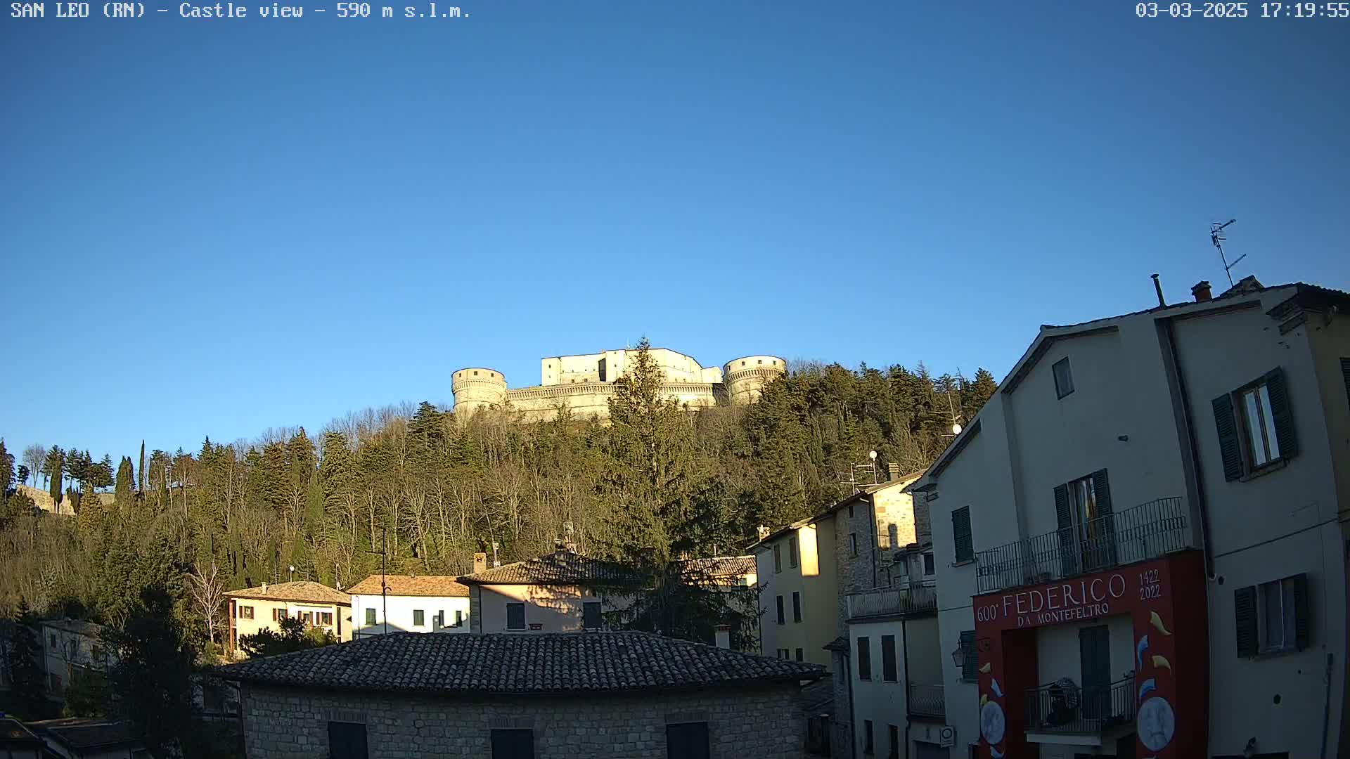 A large stone castle sits atop a hill covered in leafless trees under a clear blue sky, overlooking a village of stone buildings.