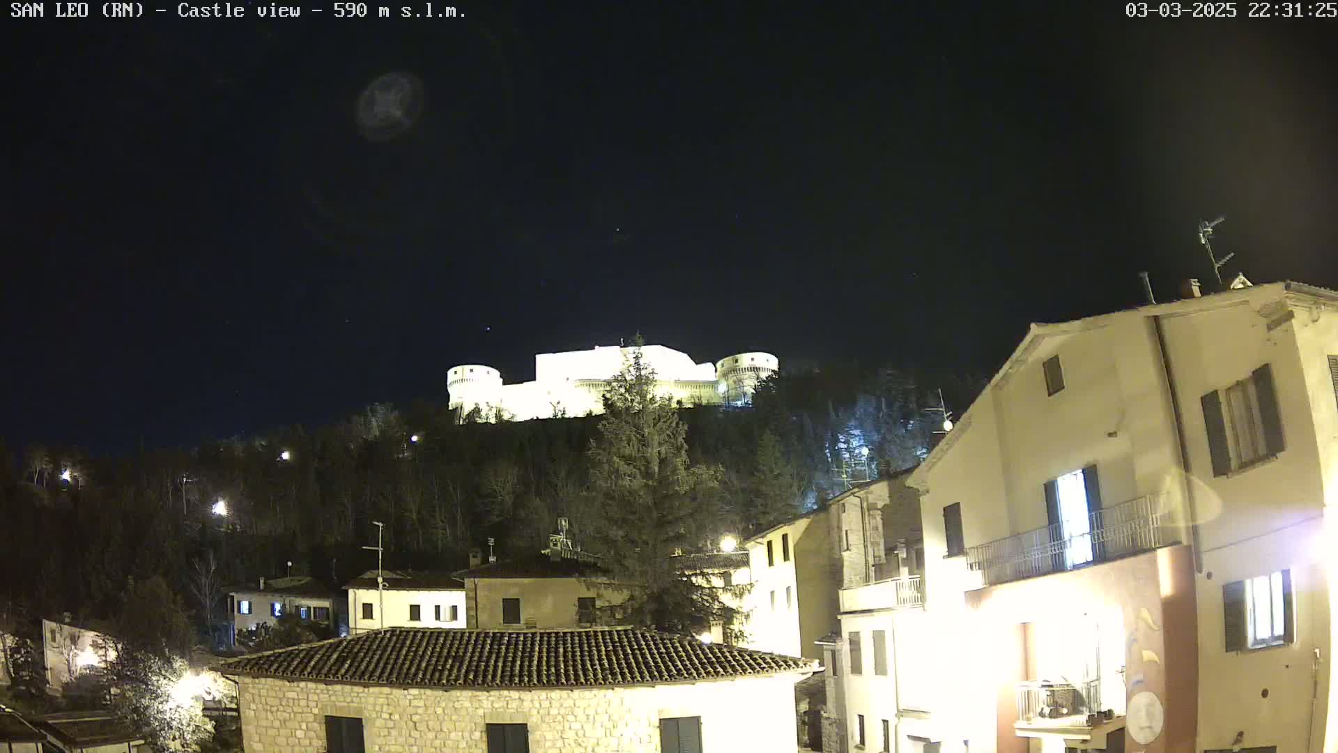 A brightly lit castle sits atop a hill overlooking a small town at night under a clear, dark sky.