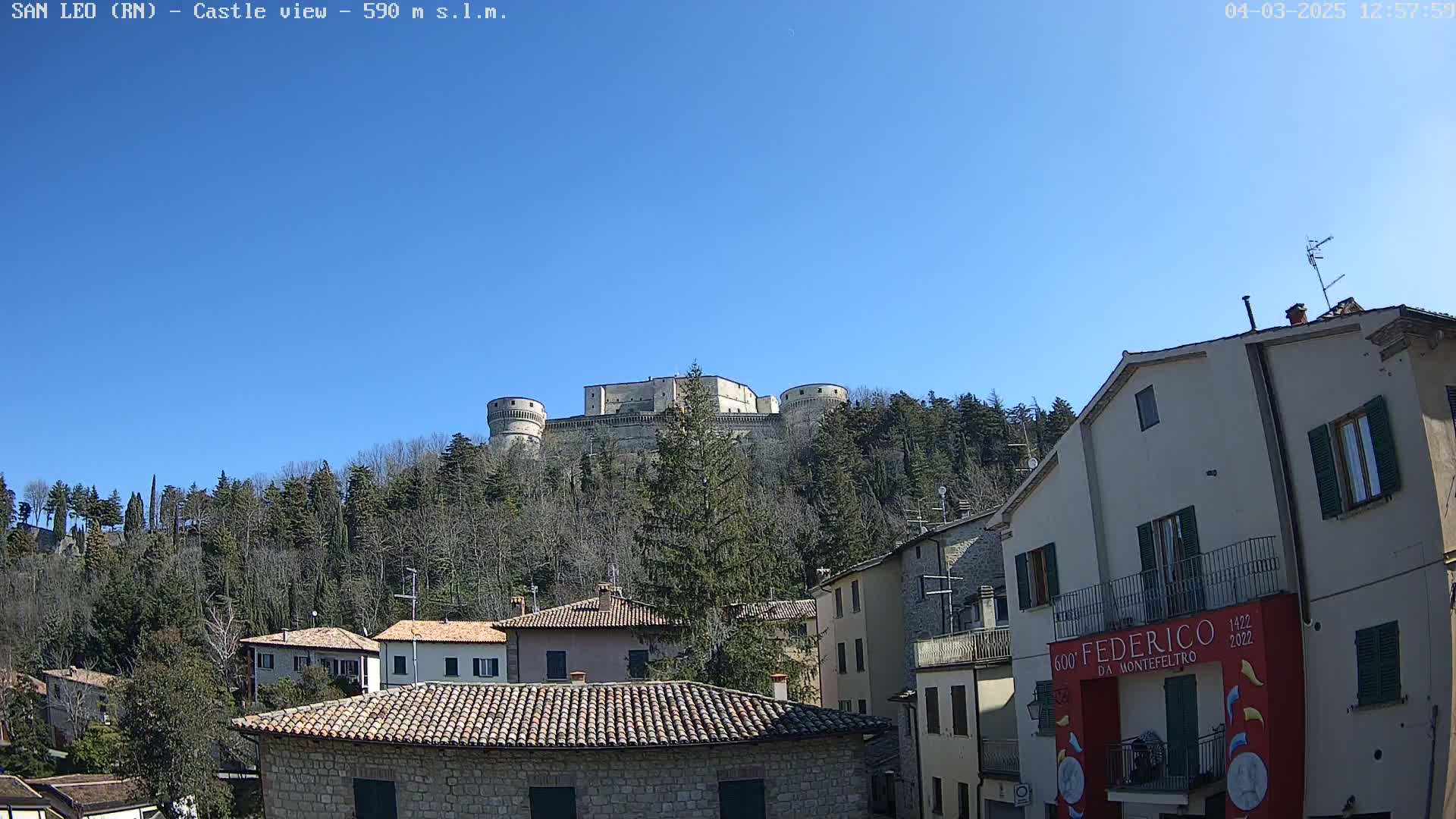 A stone castle sits atop a hill covered in leafless trees under a clear blue sky, overlooking a village of stone buildings.