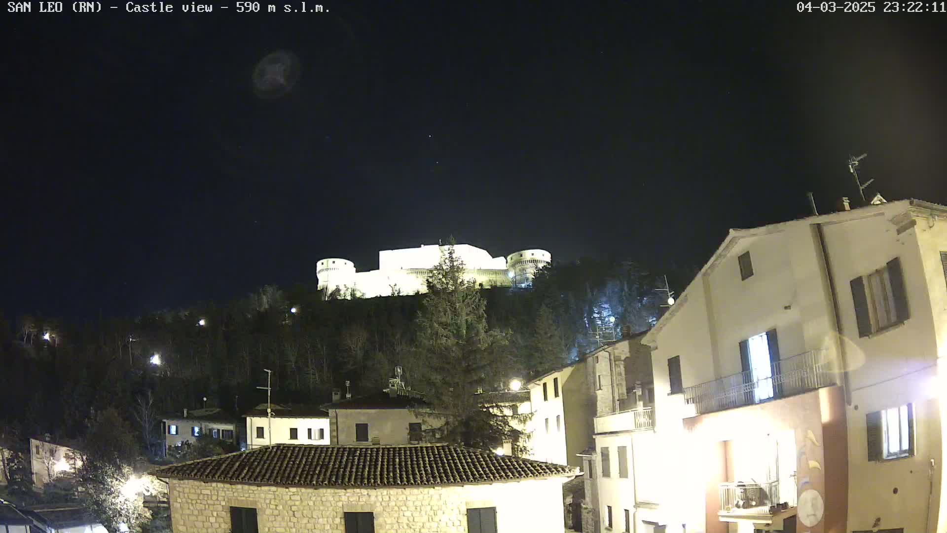 A nighttime view of a hilltop castle illuminated against a dark, clear sky, overlooking a village with buildings lit up.