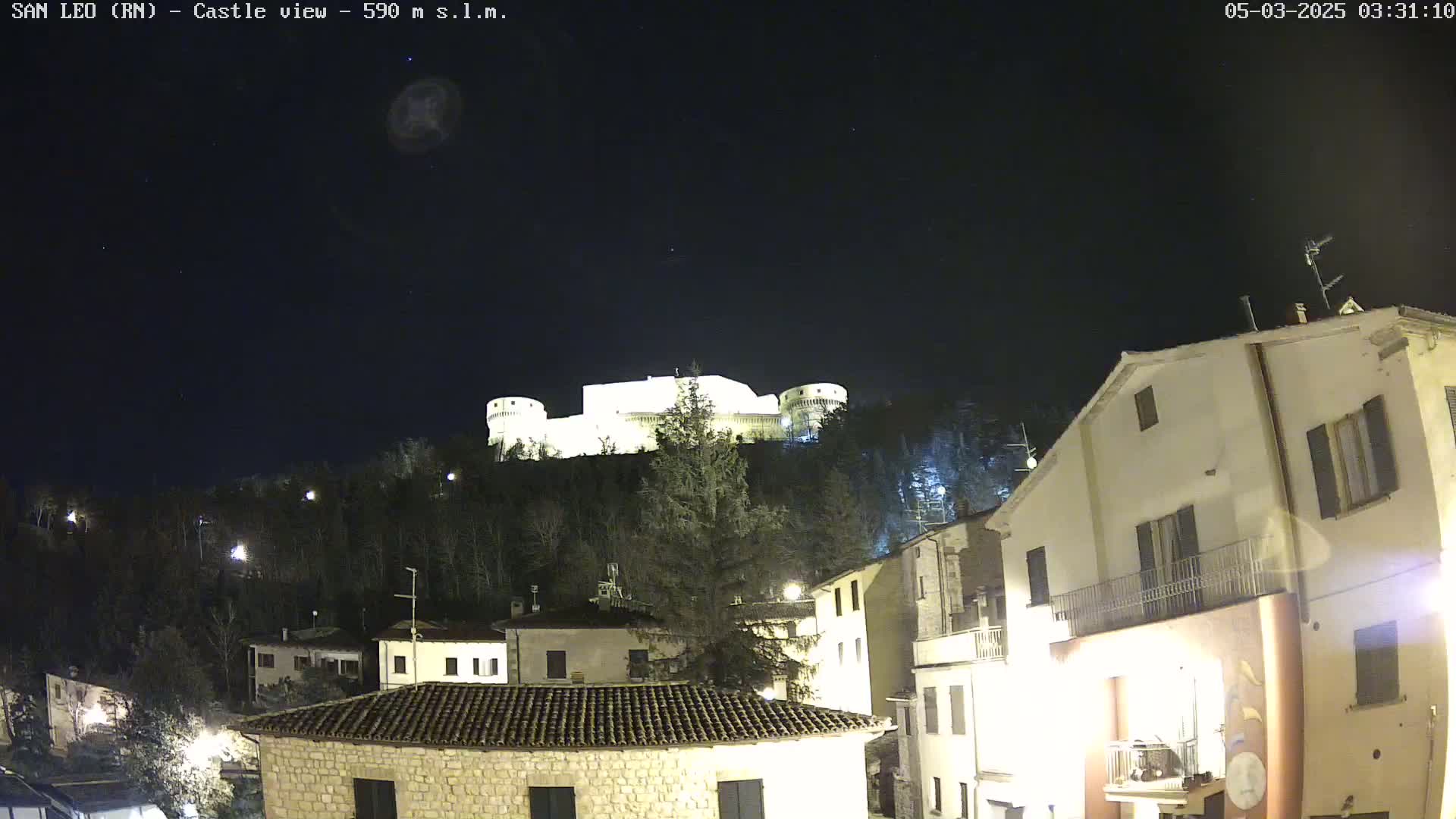 A nighttime view of a hilltop castle illuminated against a dark sky overlooking a town with buildings lit up below.