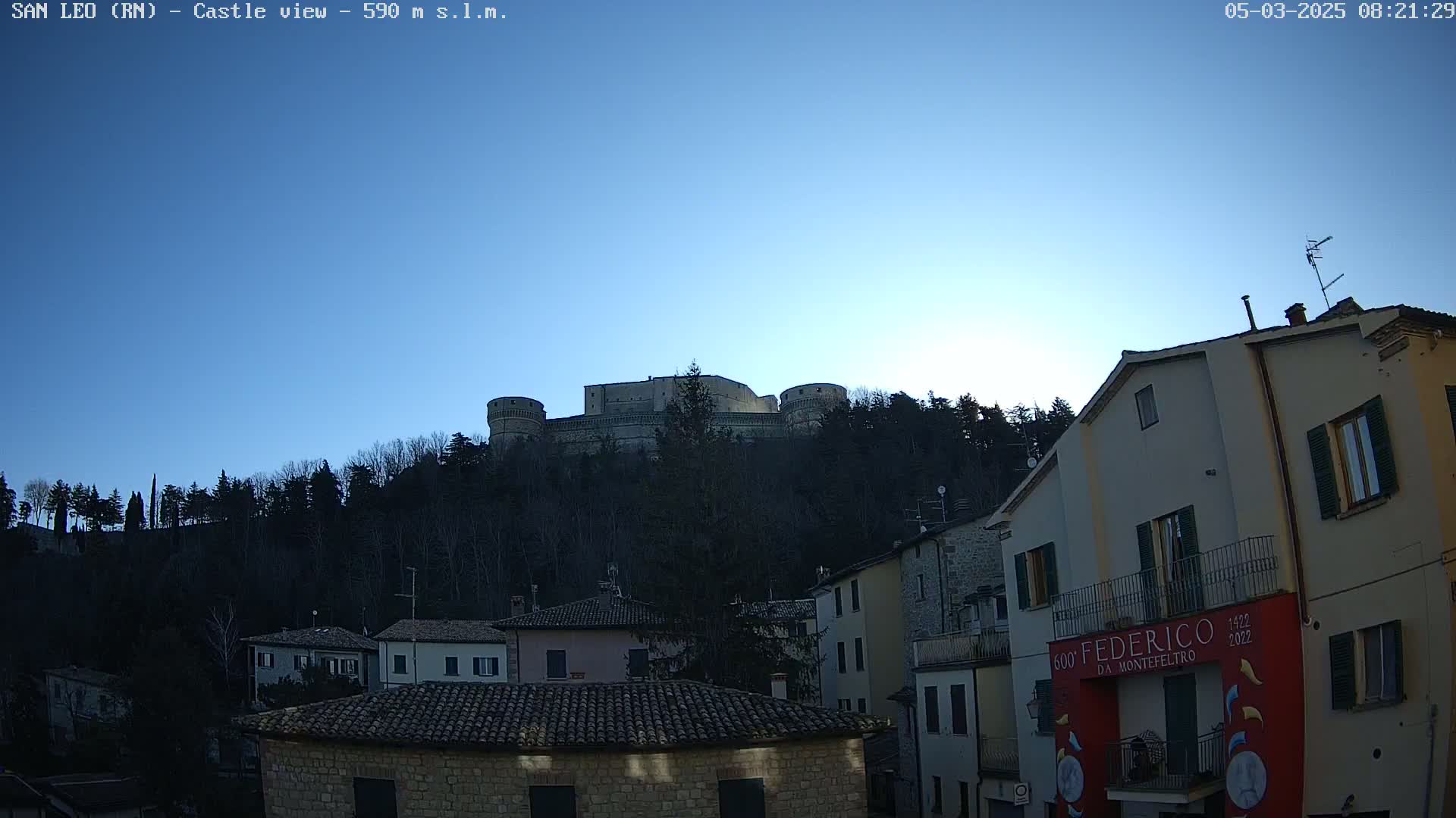 A stone castle sits atop a hill covered in leafless trees under a clear blue sky, overlooking a town with buildings of similar stone construction.
