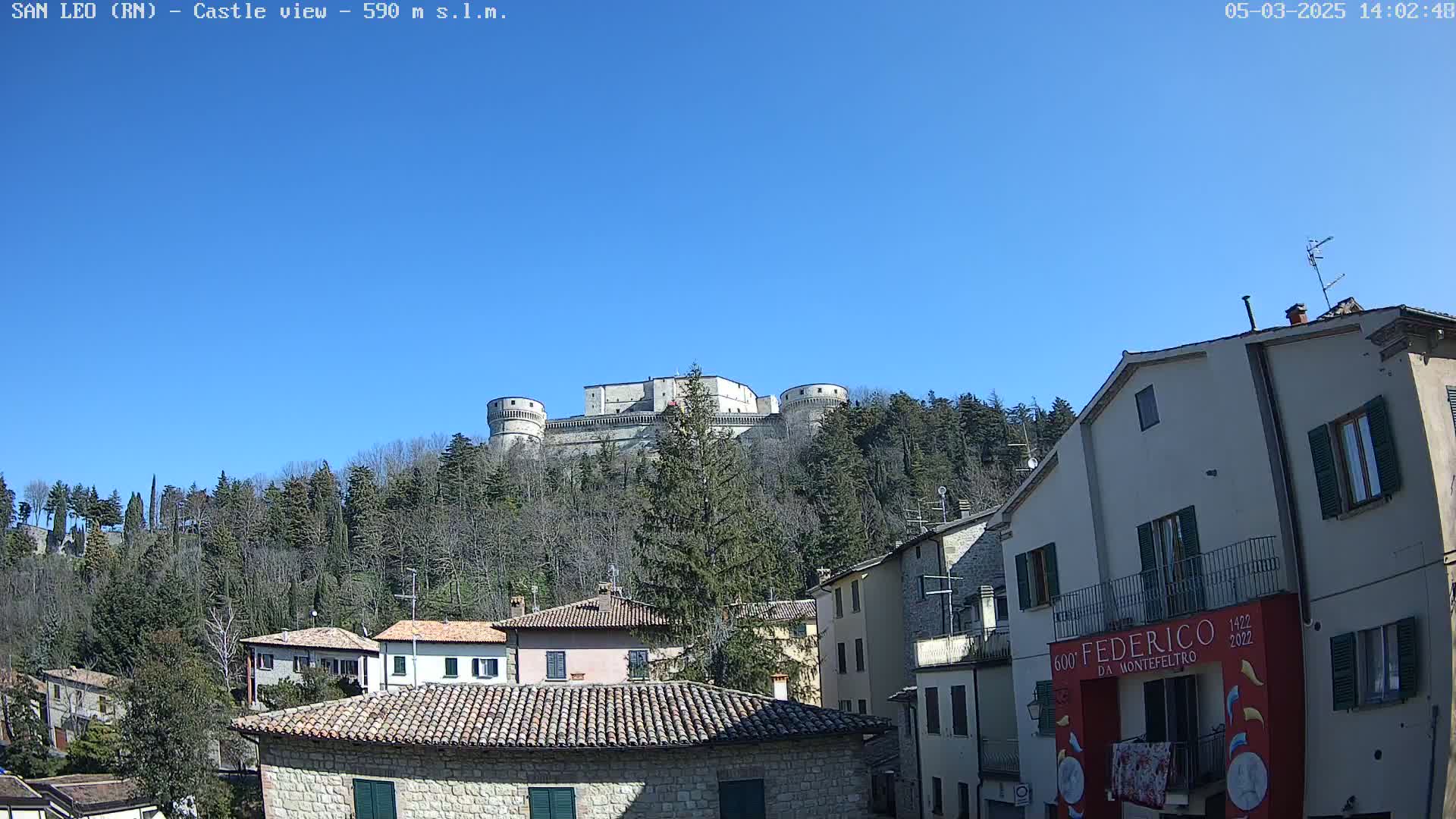 A large stone castle sits atop a hill covered in leafless trees under a clear blue sky, with a village of stone buildings in the foreground.