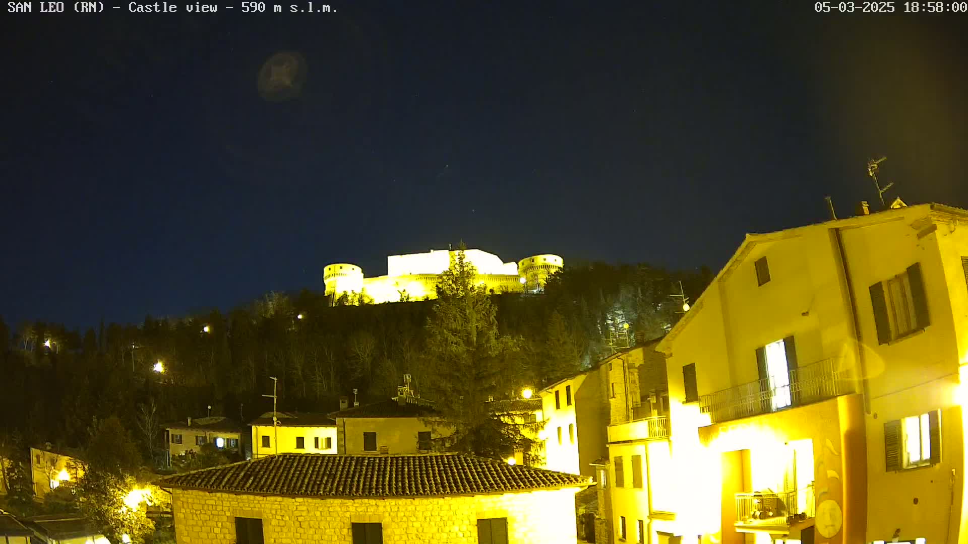 A nighttime view of a hilltop castle illuminated against a dark, clear sky, overlooking a village with buildings lit up below.