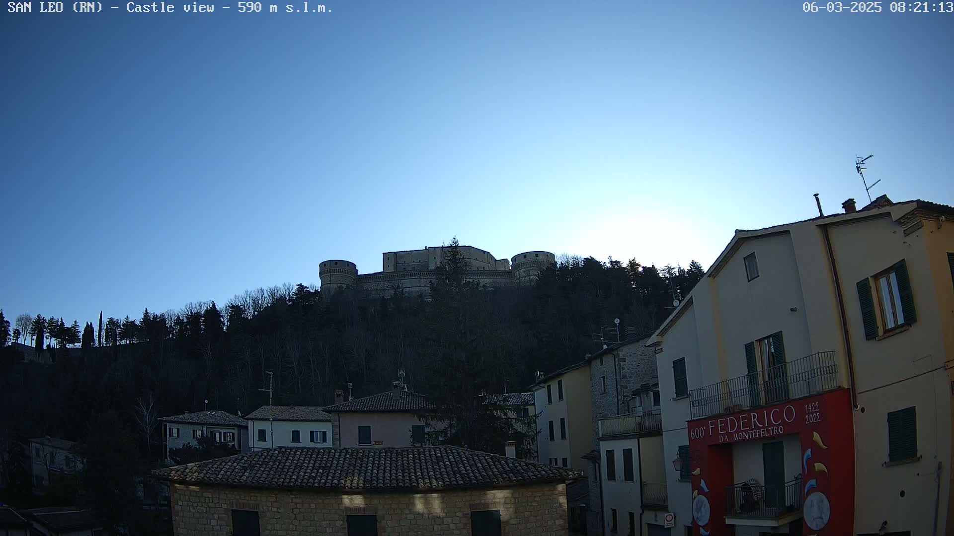 A castle sits atop a hill overlooking a town of buildings with terracotta roofs under a clear, blue sky.
