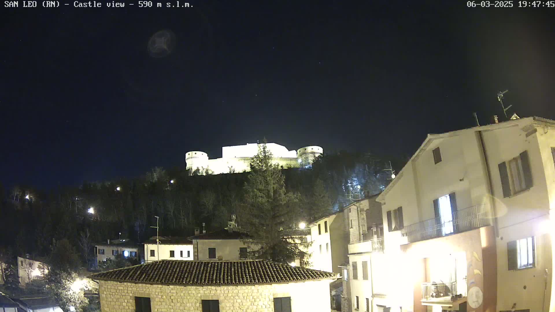 A brightly lit castle sits atop a hill at night, overlooking a town with buildings illuminated under a clear, dark sky.