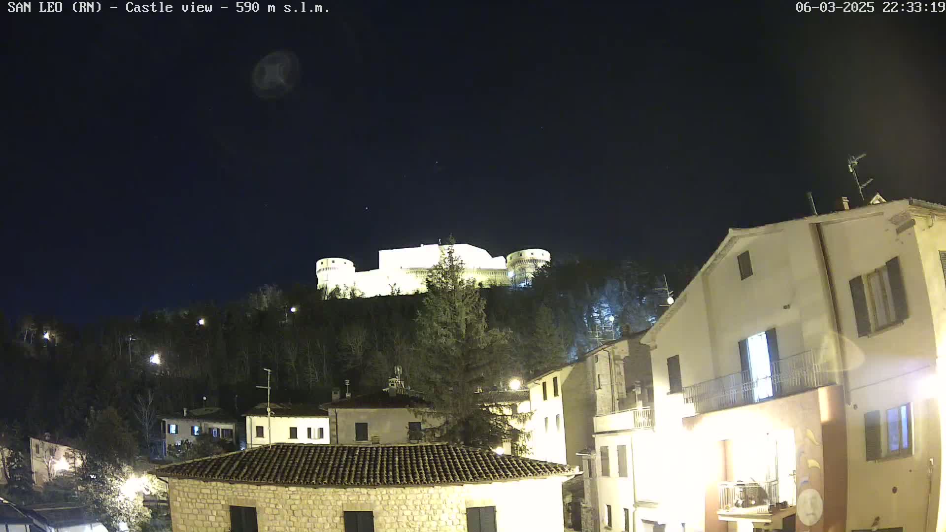 A brightly lit castle sits atop a hill overlooking a town at night under a clear, dark sky.