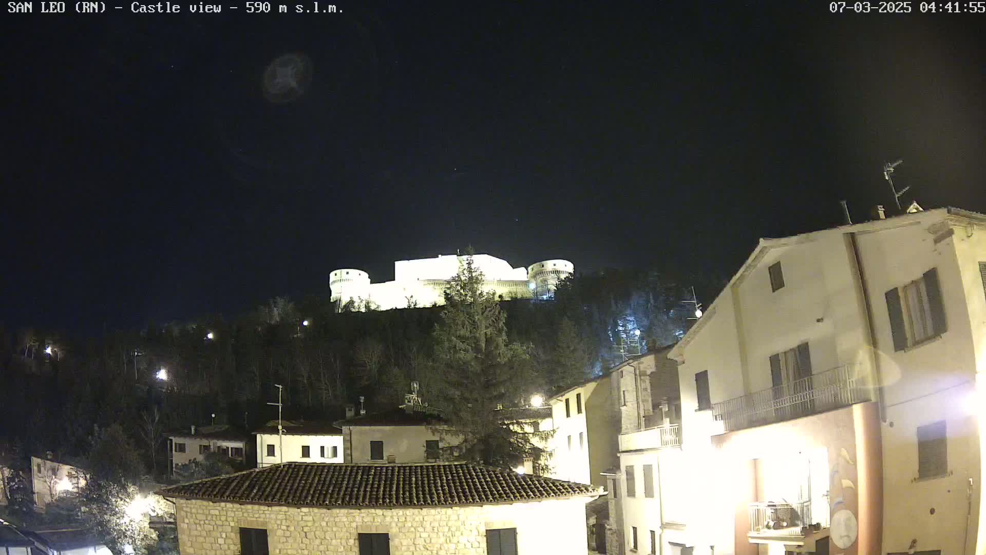 A nighttime view of a hilltop castle illuminated against a dark, clear sky, overlooking a village with buildings lit up below.