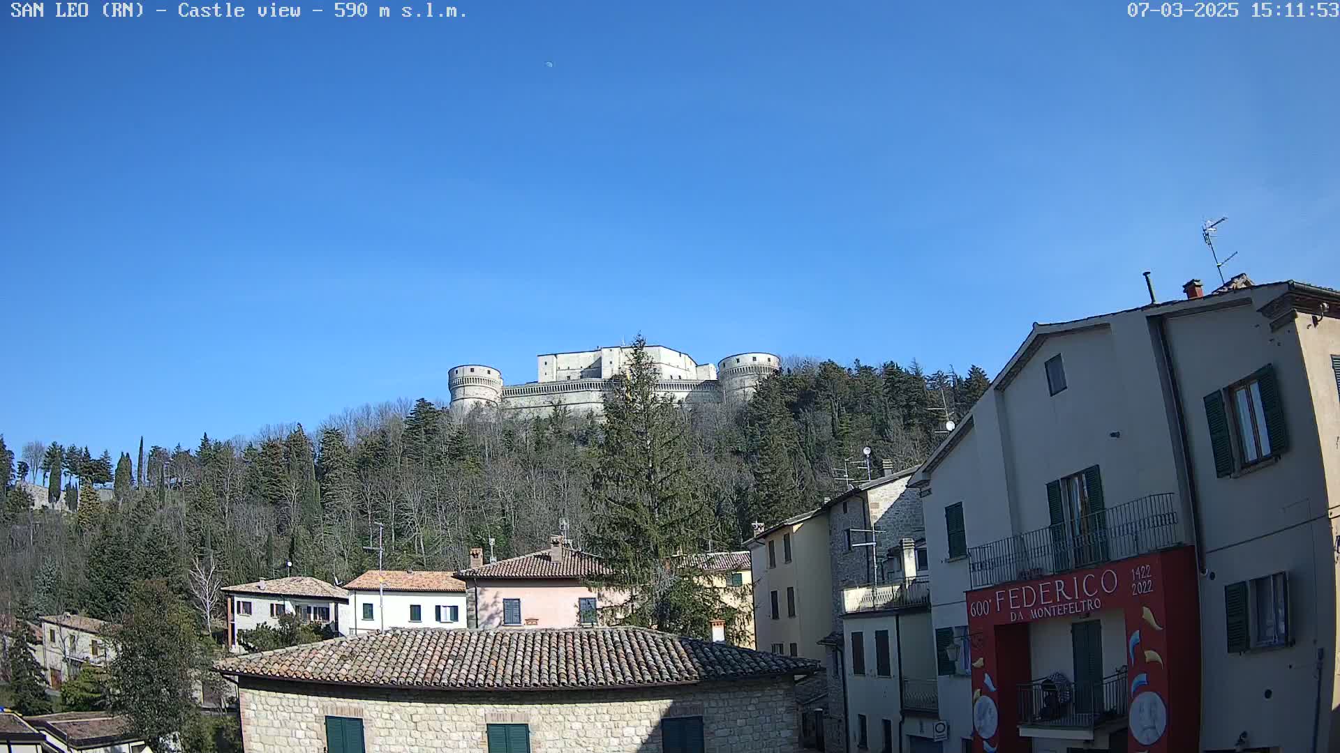 A large stone castle sits atop a hill covered in leafless trees under a clear blue sky, overlooking a village of stone buildings with terracotta roofs.
