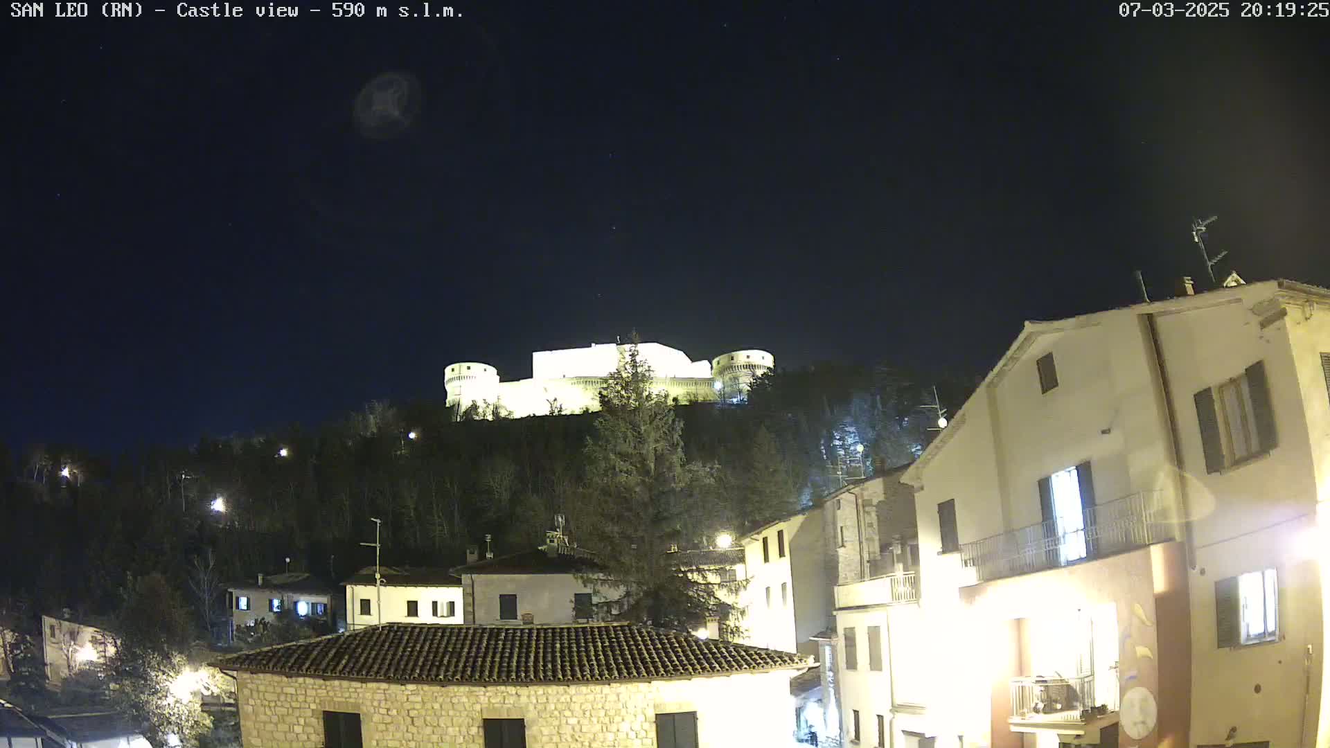A brightly lit castle sits atop a hill overlooking a town at night under a clear, dark sky.