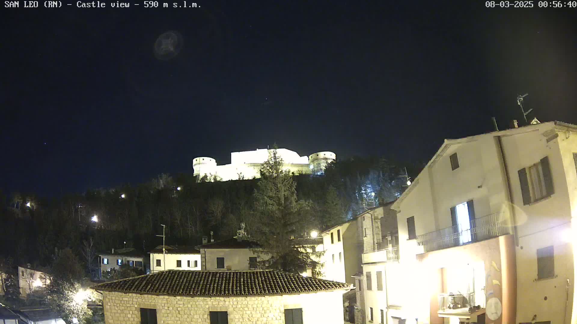 A nighttime view of a hilltop castle illuminated against a dark, clear sky, overlooking a village of low stone buildings.