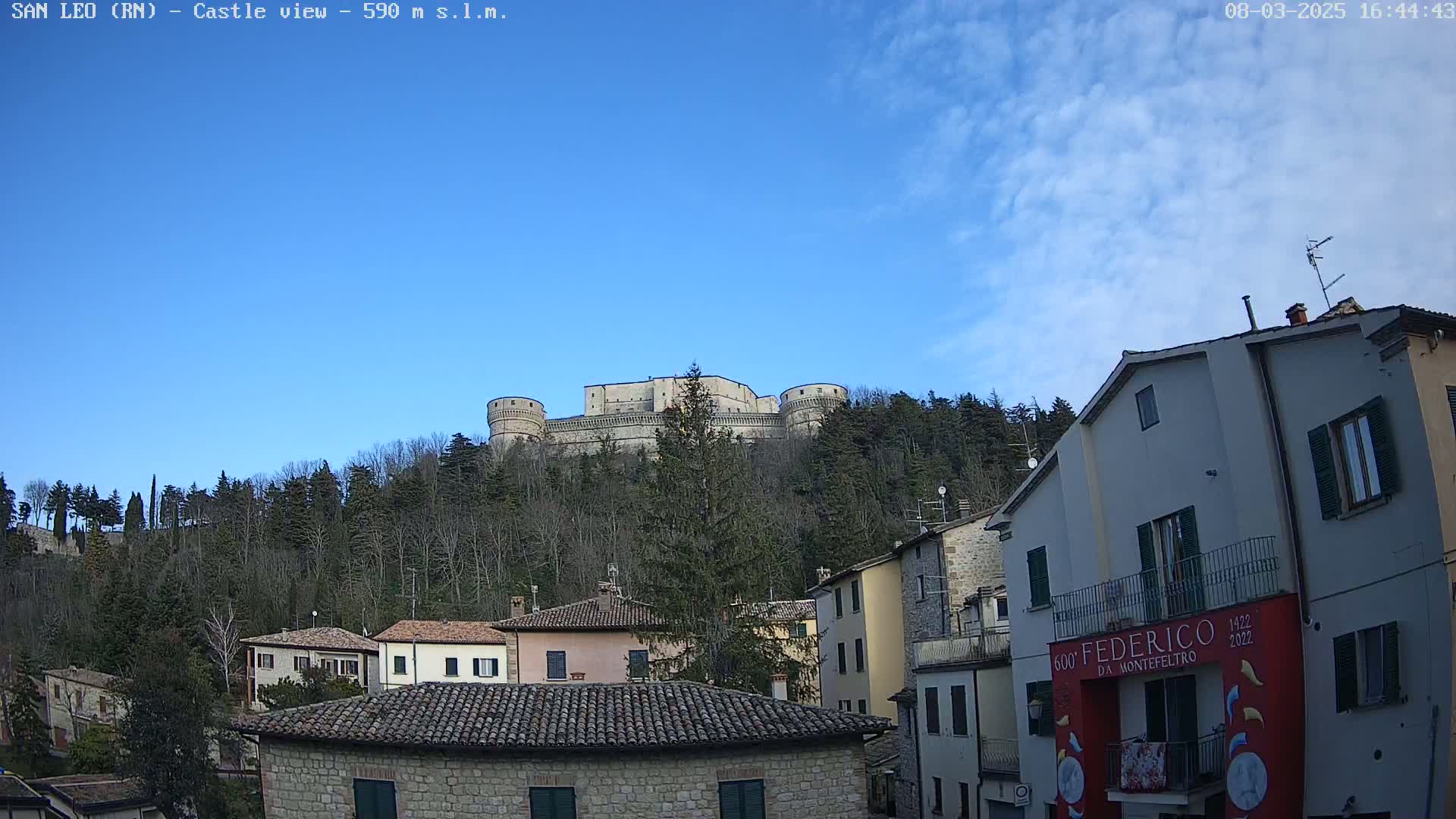 A stone castle sits atop a hill covered in leafless trees under a mostly clear blue sky with a few clouds.