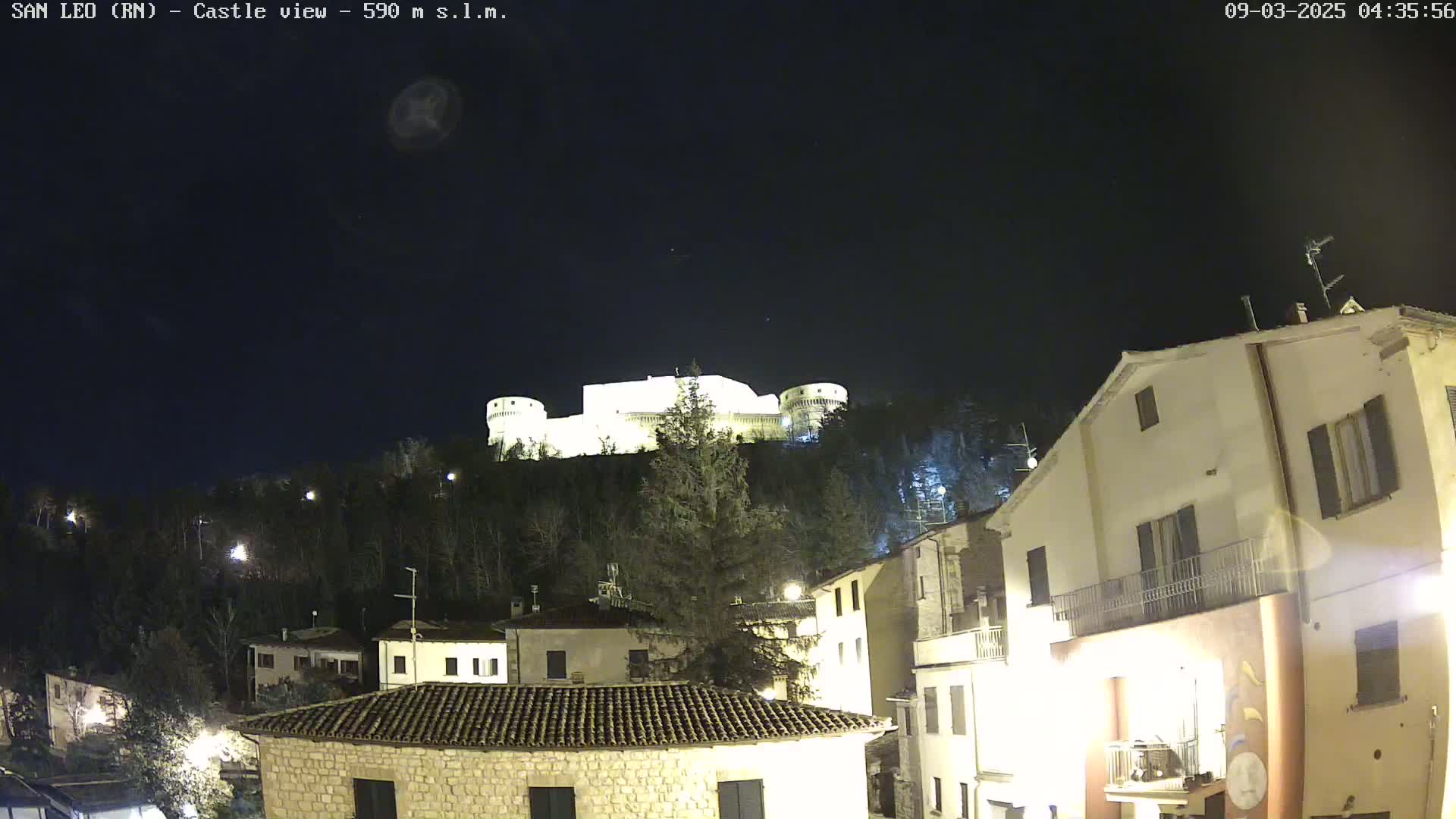 A nighttime view of a hilltop castle illuminated against a dark, clear sky, overlooking a village with buildings lit up.