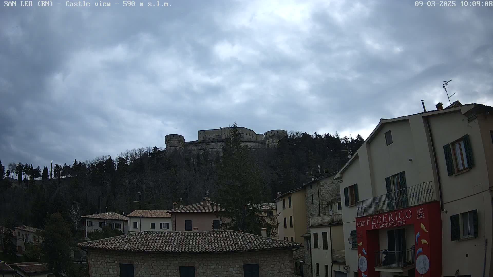 A large stone castle sits atop a hill covered in leafless trees under a cloudy sky, overlooking a village of stone buildings.