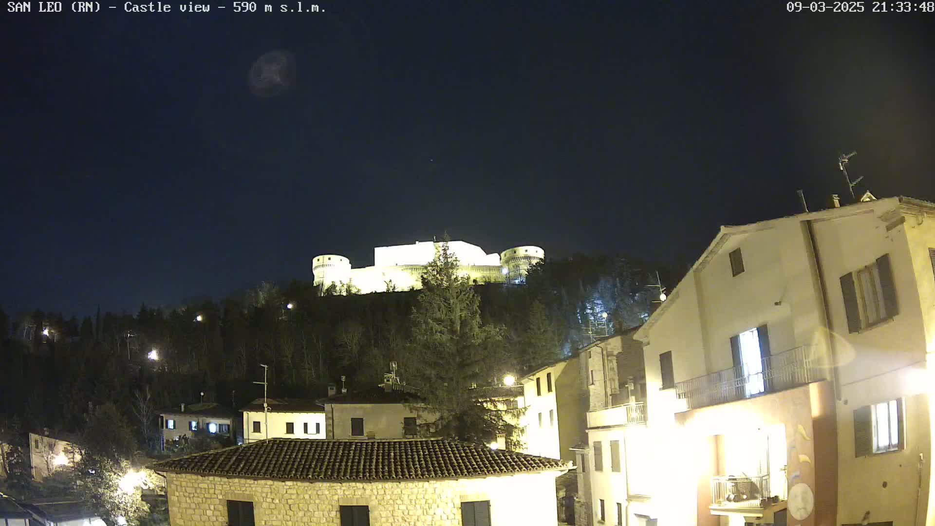 A brightly lit castle sits atop a hill at night, overlooking a town with illuminated buildings under a clear, dark sky.