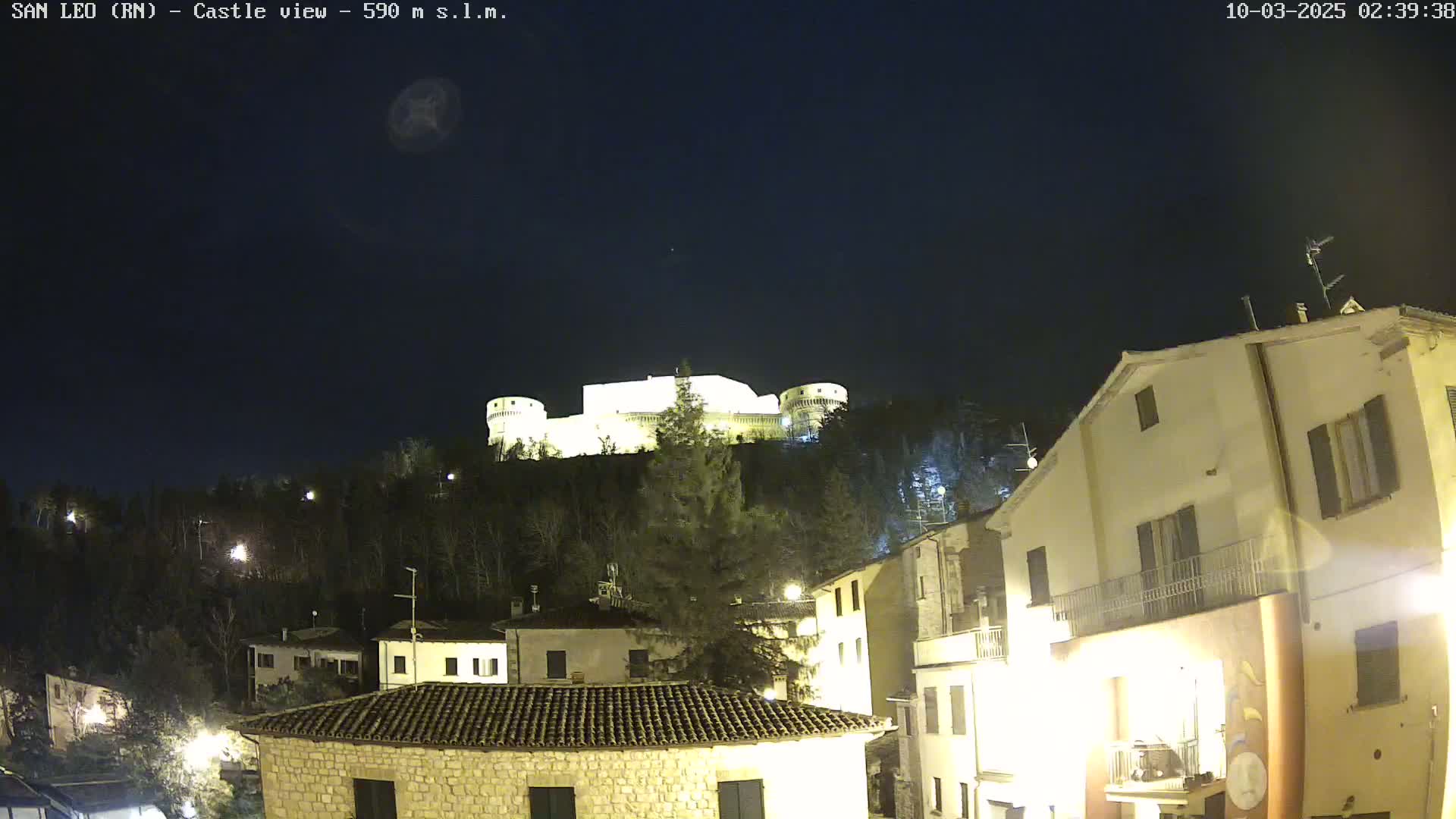 A nighttime view of a hilltop castle illuminated against a dark sky, overlooking a town with buildings lit up below.