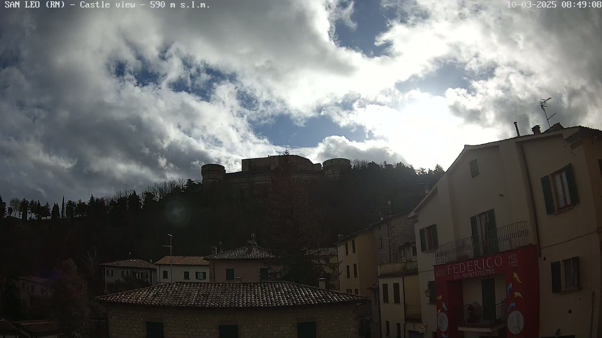 A hilltop castle is visible through a partly cloudy sky above a town of low stone buildings.