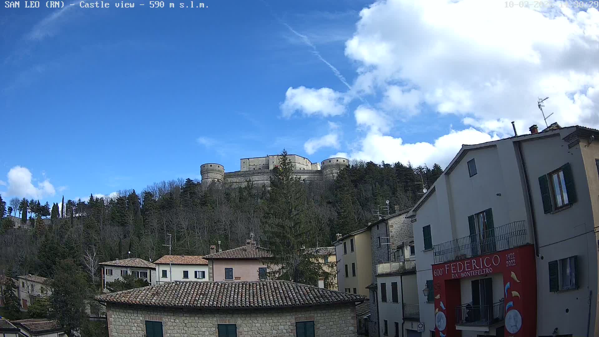 A stone castle sits atop a hill covered in leafless trees under a partly cloudy blue sky, overlooking a town of stone buildings.
