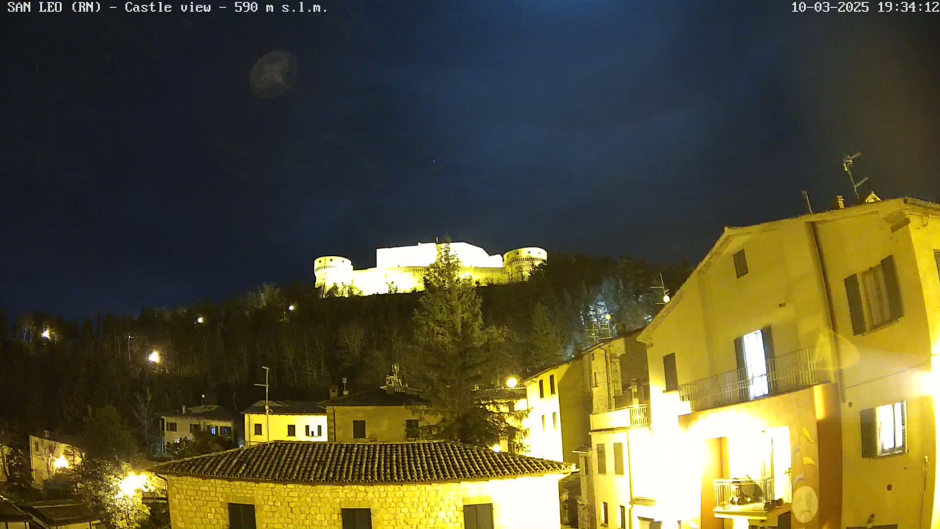 A nighttime view of a hilltop castle illuminated against a dark, clear sky, overlooking a village with buildings lit up below.