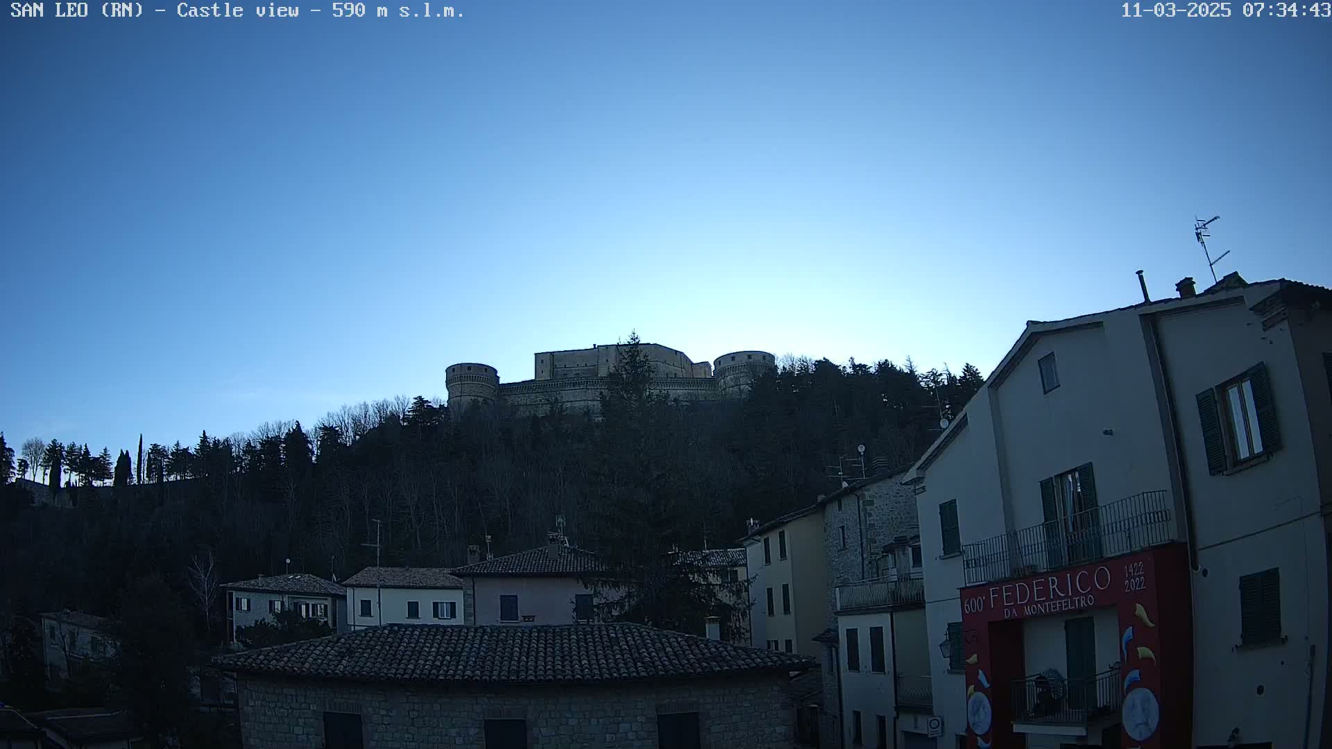 A castle sits atop a hill covered in dark trees under a clear, twilight sky, overlooked by a town of stone buildings.