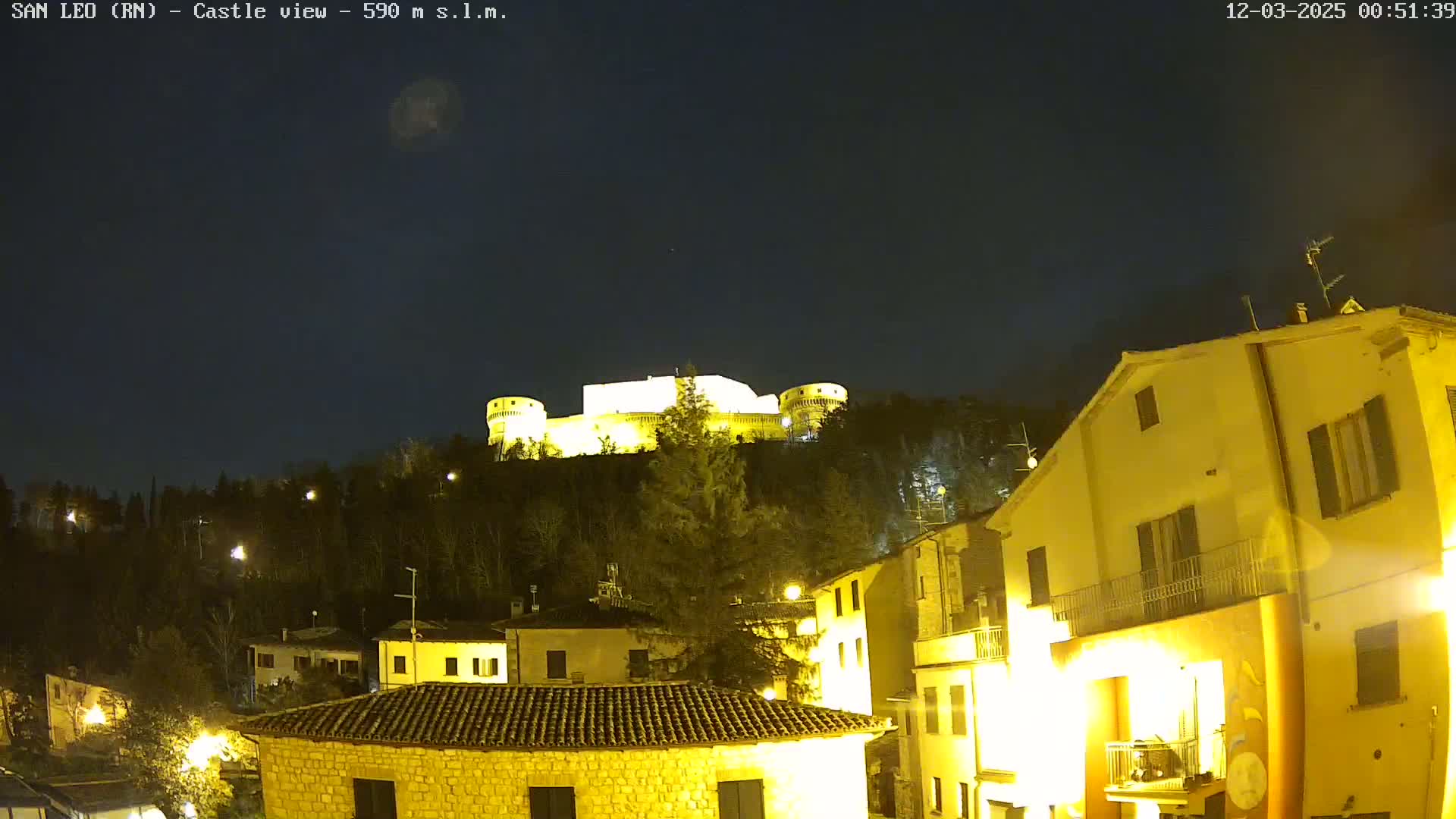 A nighttime view of a hilltop castle illuminated against a dark, clear sky, overlooking a village with buildings lit up below.