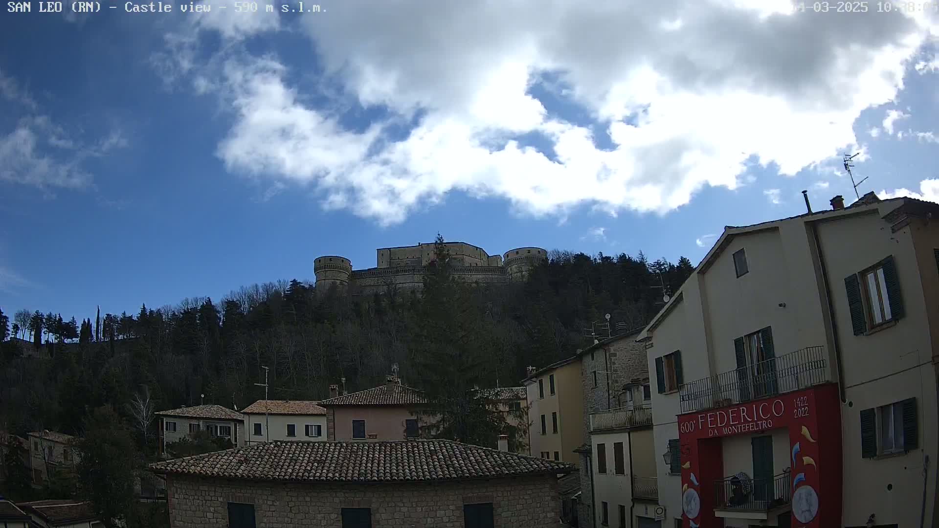 A large stone castle sits atop a hill covered in leafless trees under a partly cloudy sky, overlooking a village of stone buildings.
