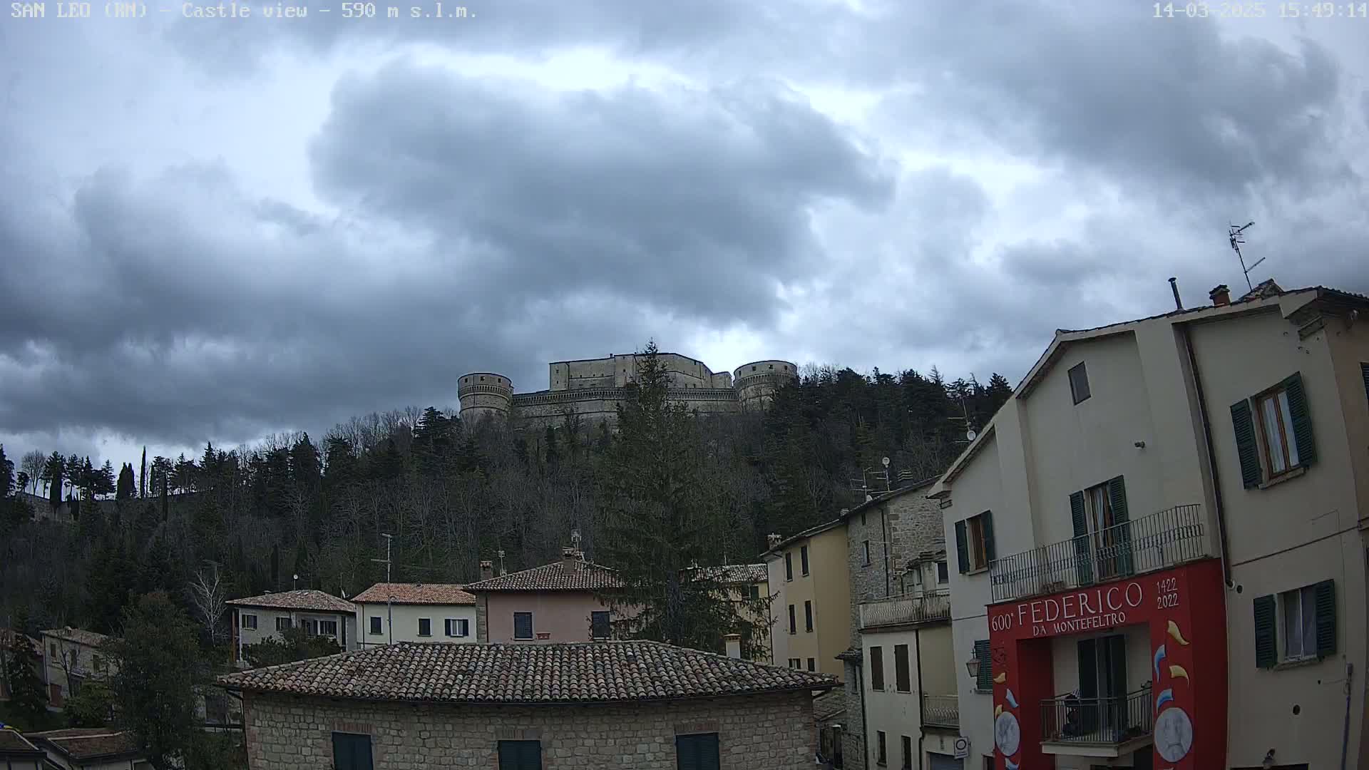 A stone castle sits atop a hill covered in leafless trees under a cloudy sky, overlooking a town of stone buildings with red tile roofs.