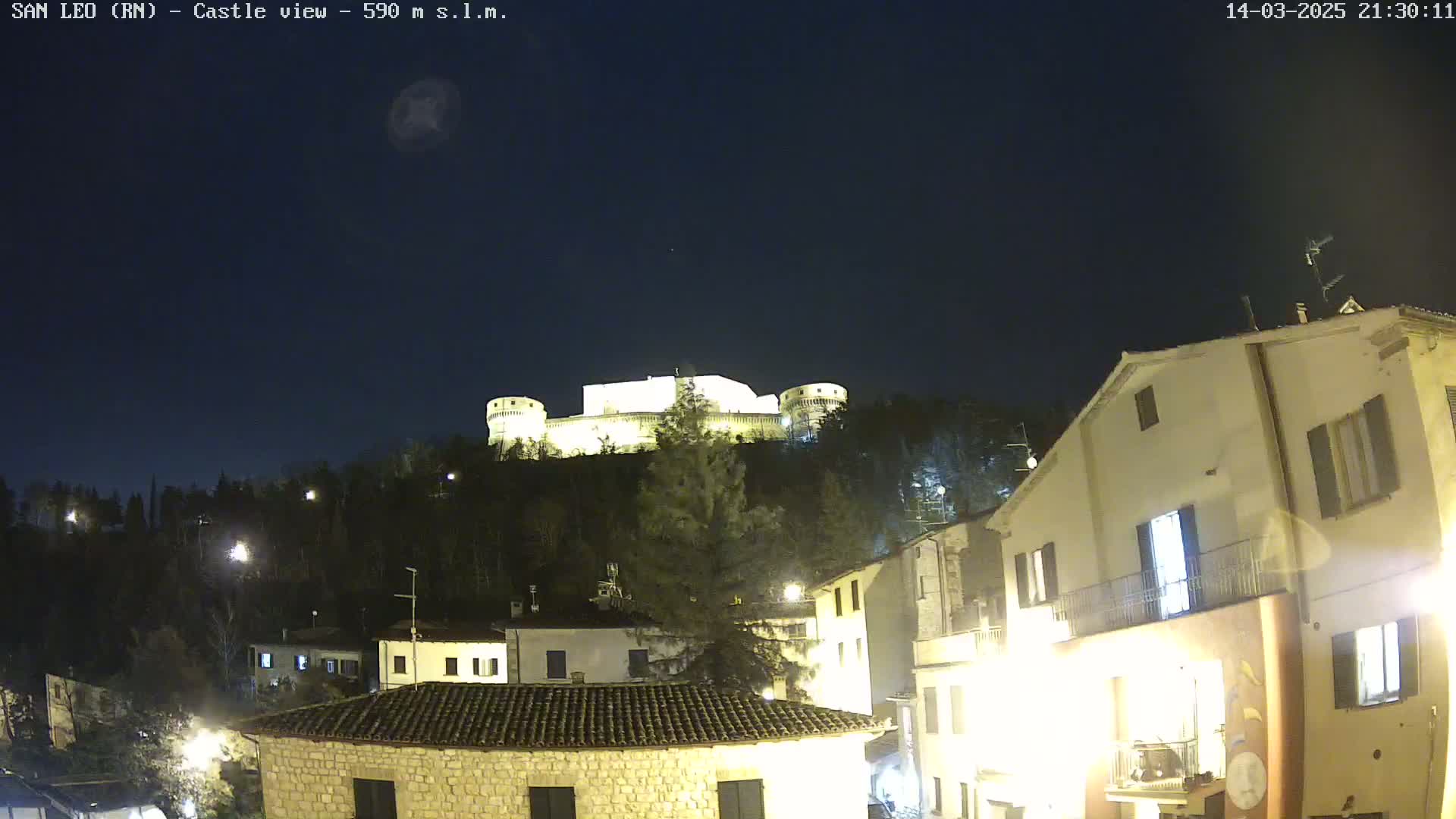 A nighttime view of a hilltop castle illuminated against a dark, clear sky, with a village of buildings at the base of the hill.