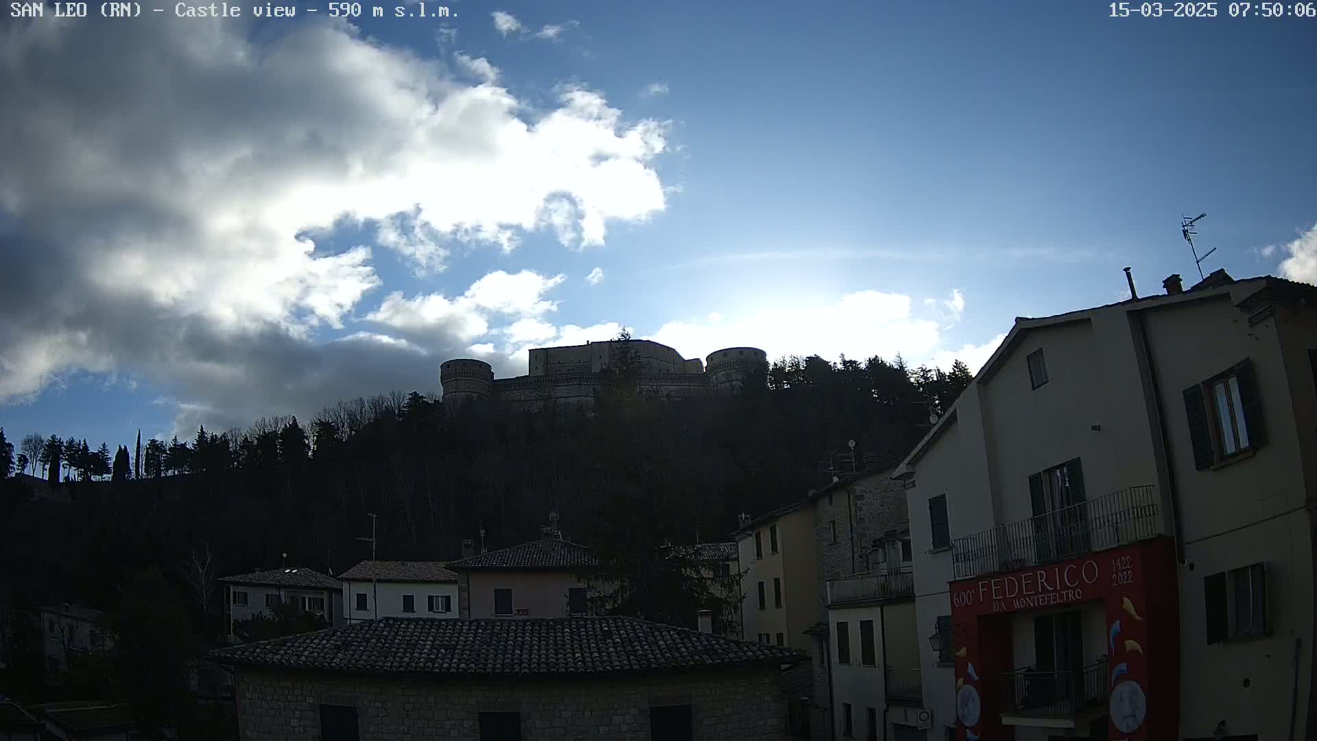 A partly cloudy sky overlooks a hilltop castle, visible behind a row of buildings.
