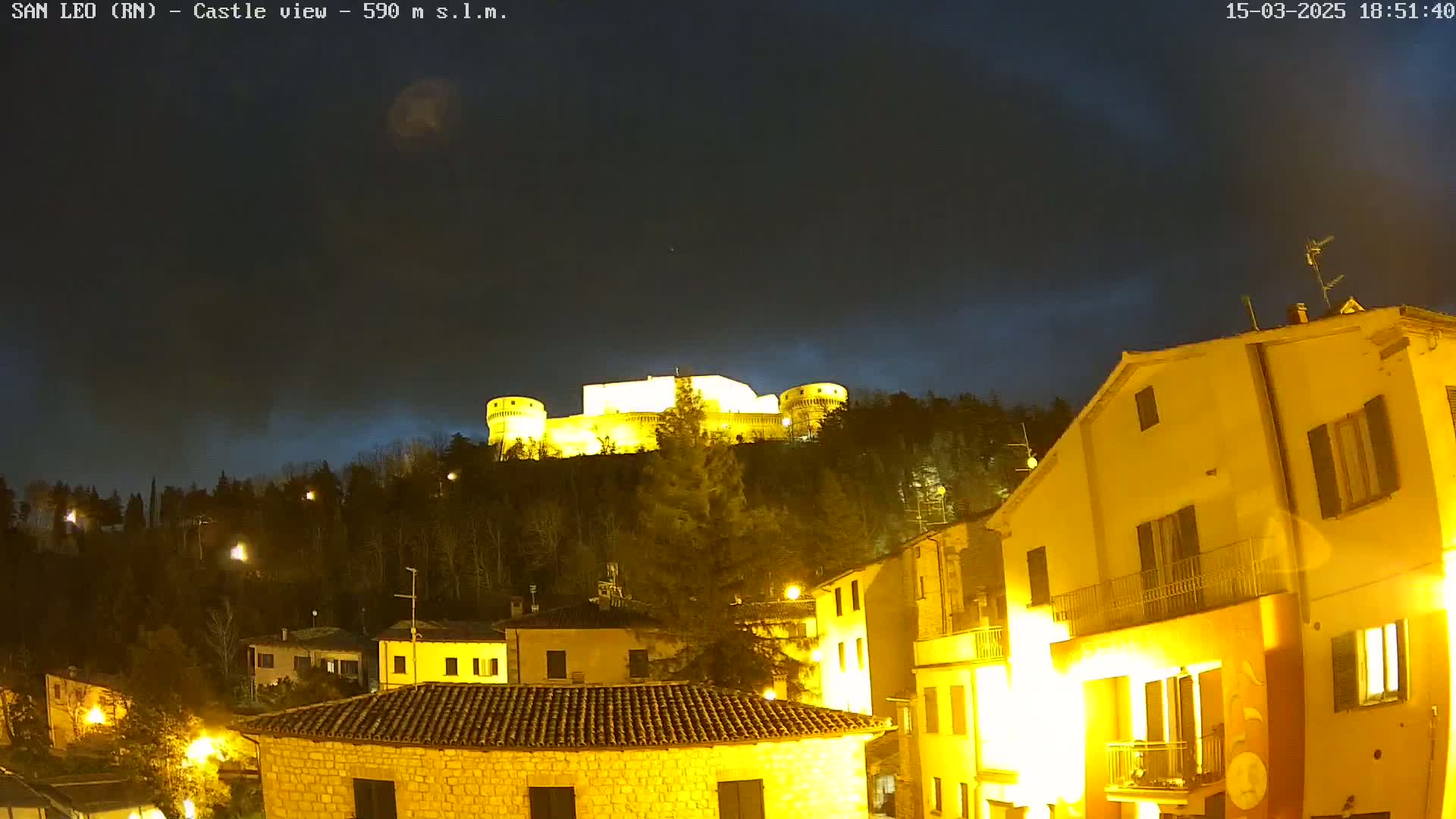 A brightly lit castle sits atop a hill at night, overlooking a town with illuminated buildings under a mostly clear, dark sky.