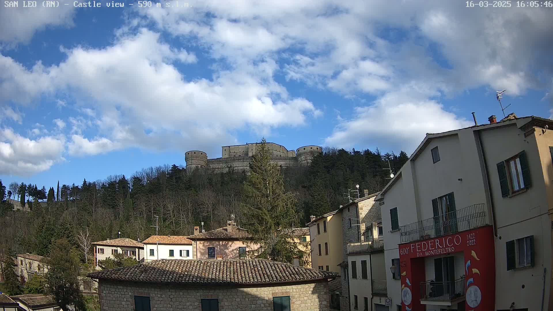 A large stone castle sits atop a hill covered in leafless trees under a partly cloudy sky.