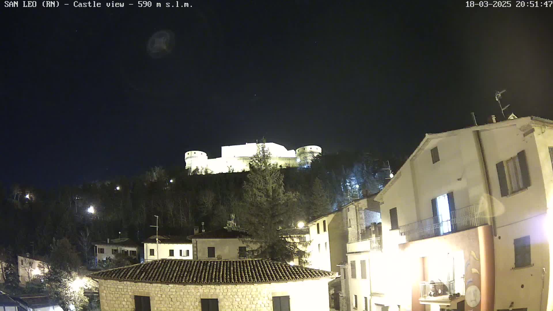 A brightly lit castle sits atop a hill at night, overlooking a village with buildings illuminated by streetlights under a clear, dark sky.