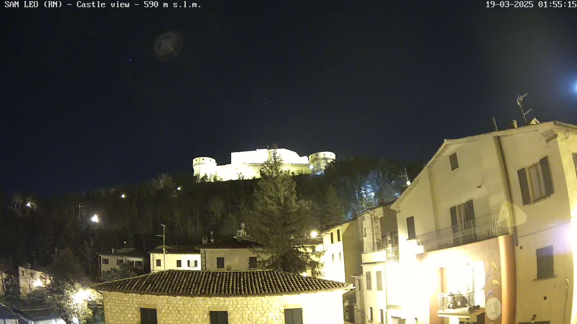 A brightly lit castle sits atop a hill at night, overlooking a village of illuminated buildings under a clear, starry sky.