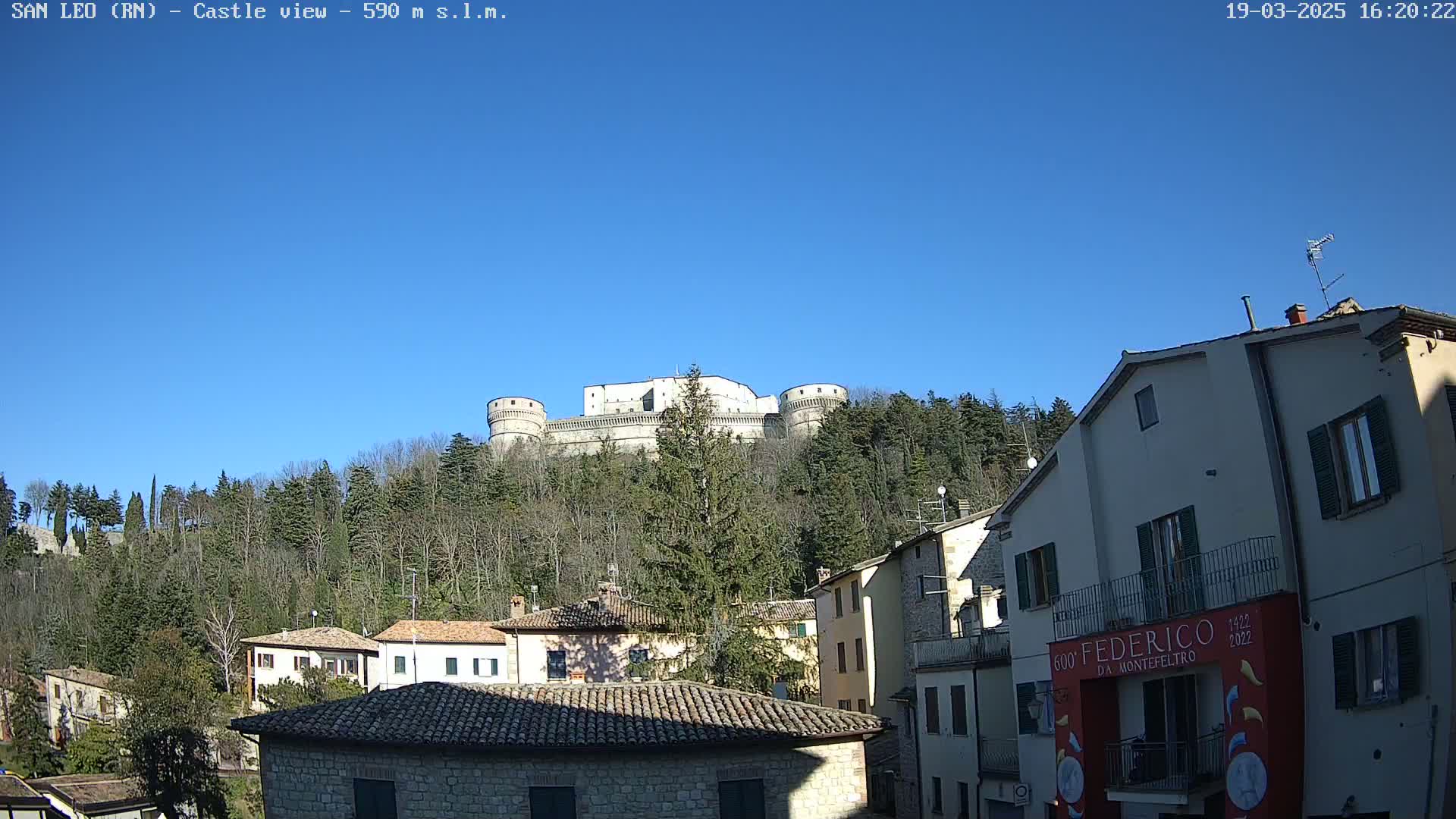 A large stone castle sits atop a hill covered in trees under a clear blue sky, overlooking a town of smaller stone buildings.