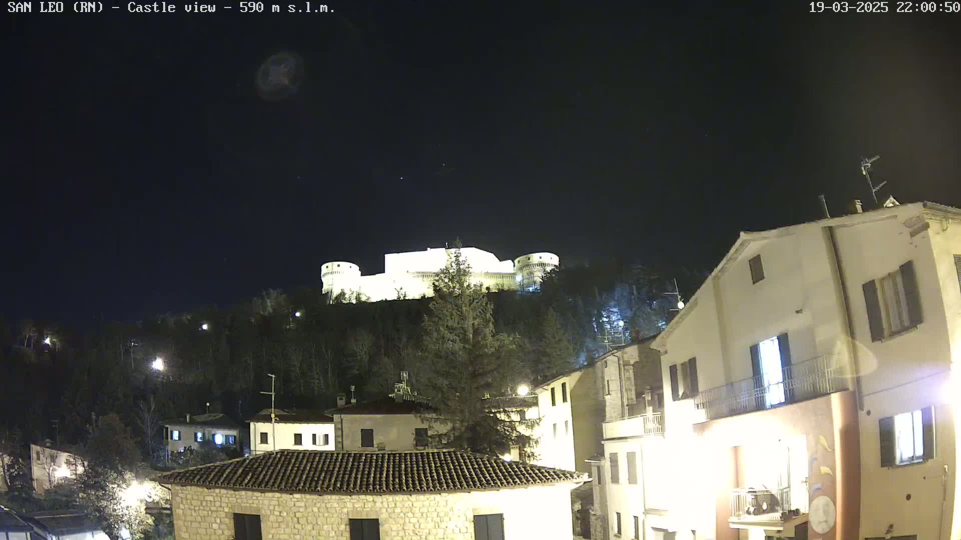 A brightly lit castle sits atop a hill overlooking a village at night under a clear, dark sky.