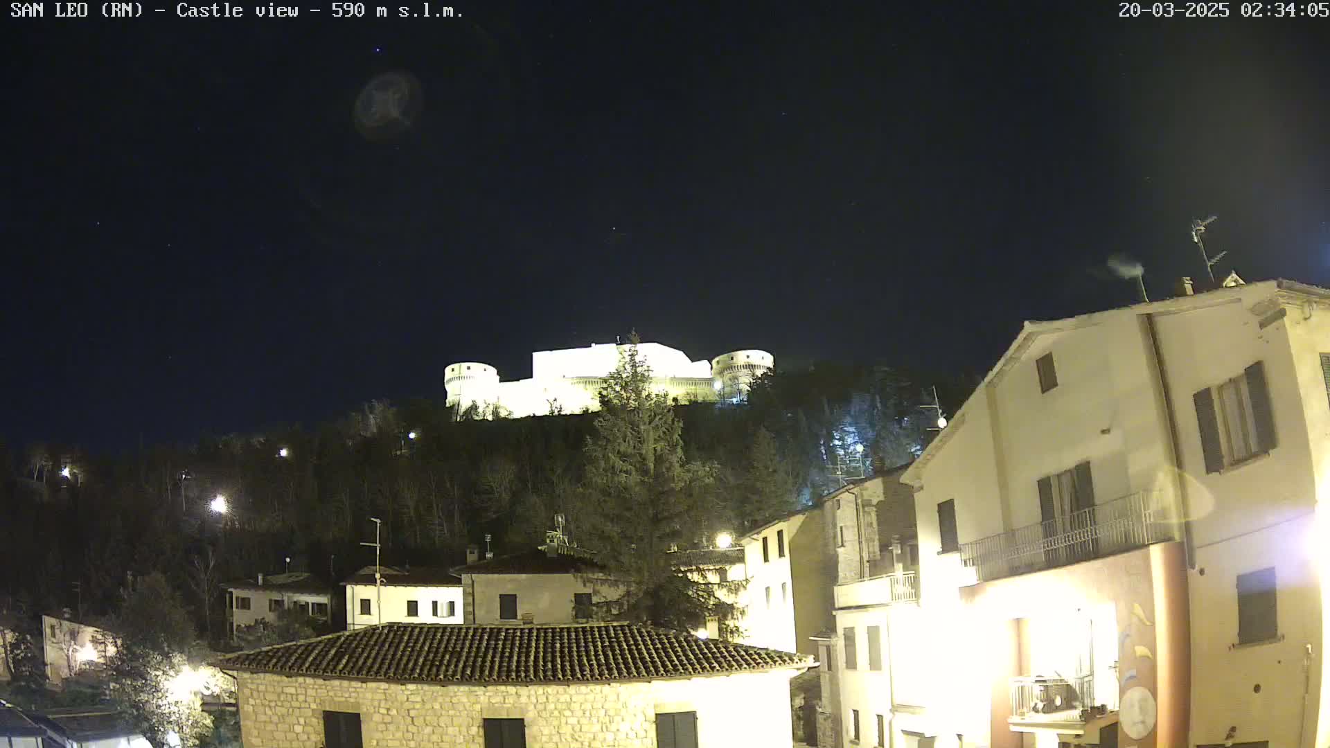 A brightly lit castle sits atop a hill at night, overlooking a village with several buildings under a clear, dark sky.