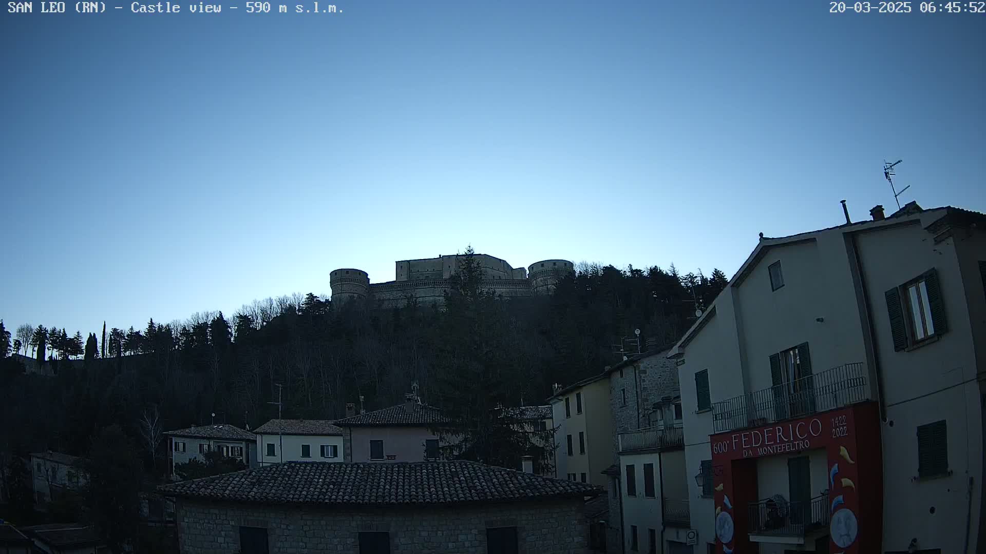A large stone castle sits atop a hill covered in dark trees under a clear, twilight sky, overlooking a village of closely spaced buildings.