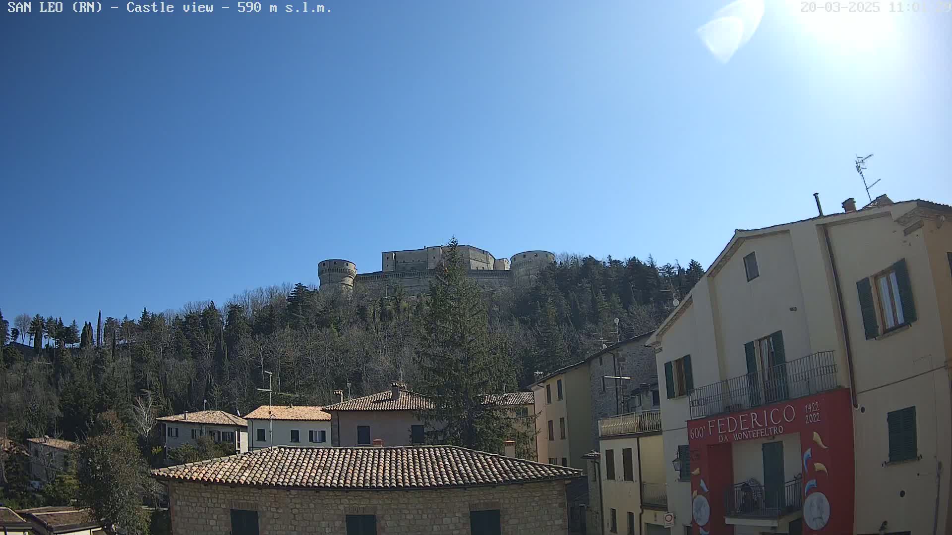 A stone castle sits atop a hill covered in leafless trees under a clear blue sky, overlooking a town with terracotta-roofed buildings.