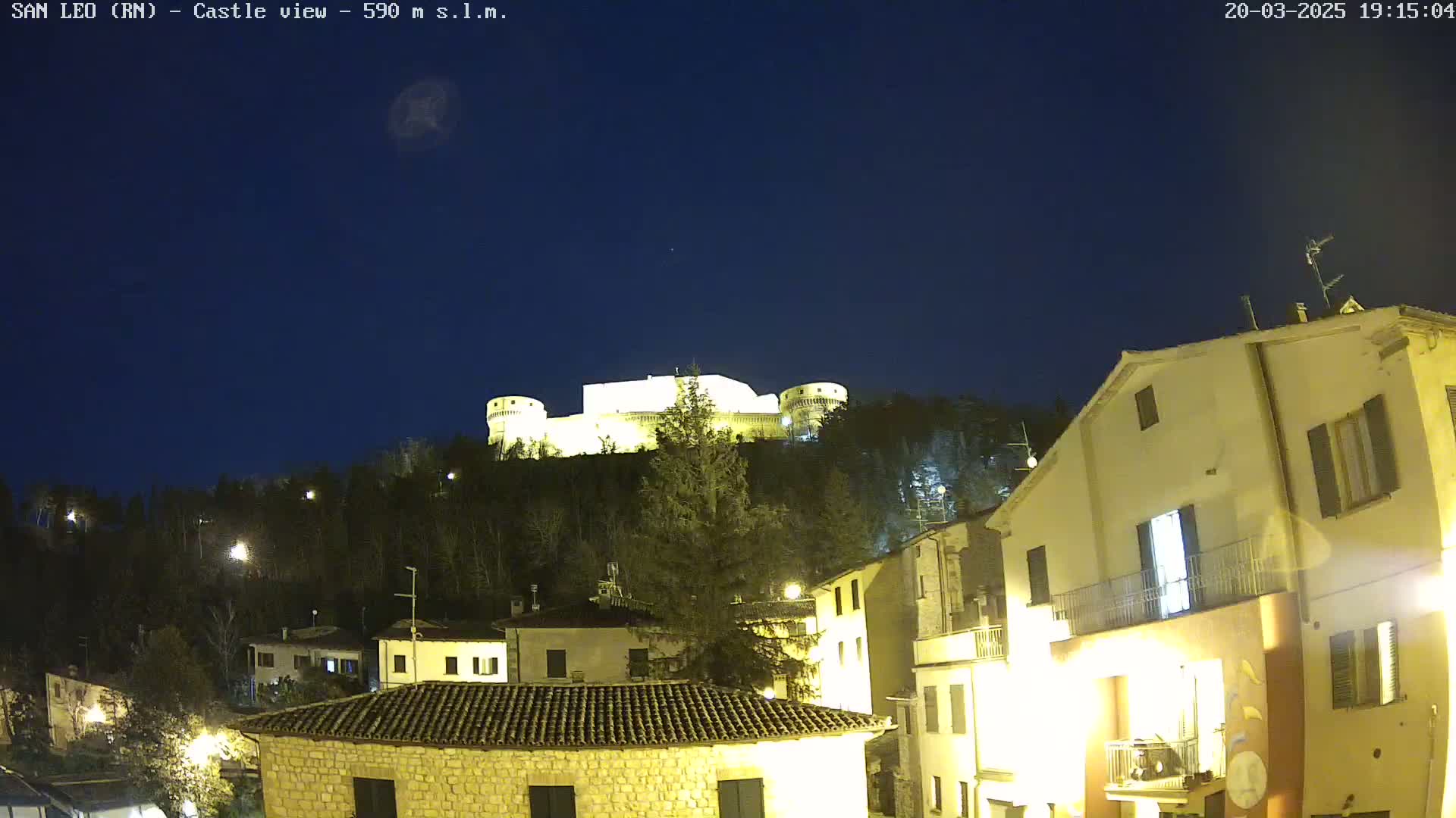 A nighttime view of a hilltop castle illuminated, overlooking a village with buildings lit up under a clear, dark sky.